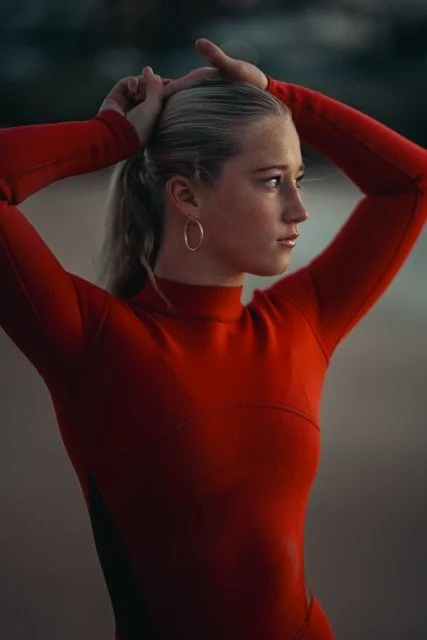 A woman with blonde hair pulled back, wearing hoop earrings and a red long-sleeve top, stands outdoors with water and distant land in the background. She is touching her head with both hands and looking to the side.