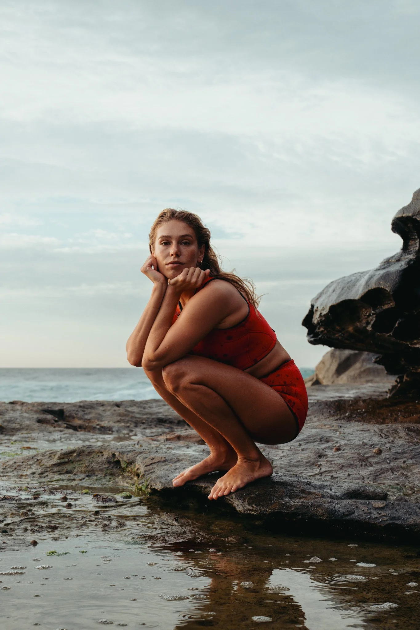 A woman crouching on a rock at the beach, wearing a red sports bra and matching shorts, with her chin resting on her hands and looking at the camera.