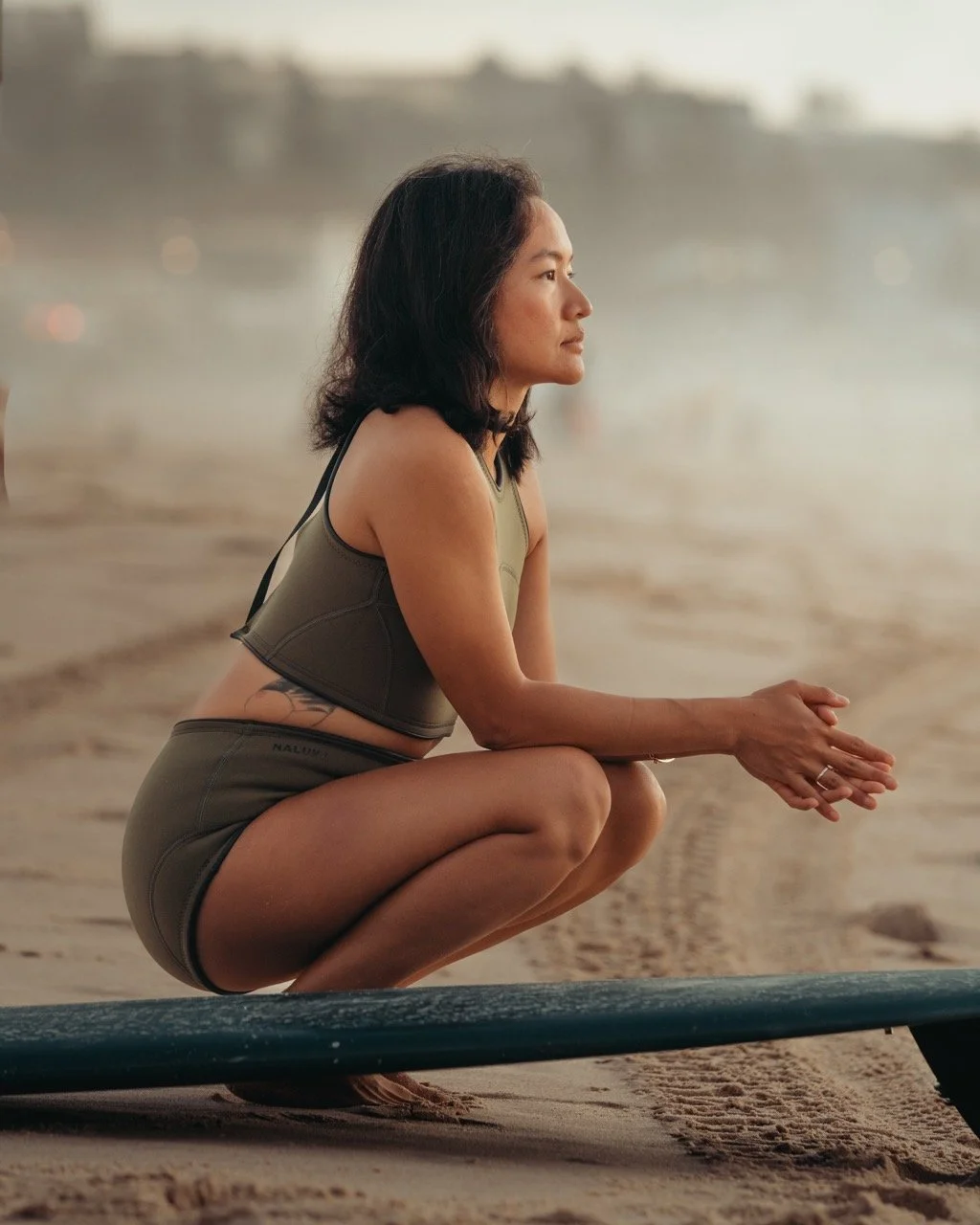 A woman crouches on a yoga mat on a sandy beach, looking contemplative as she gazes into the distance with a blurred background of the beach and sky.