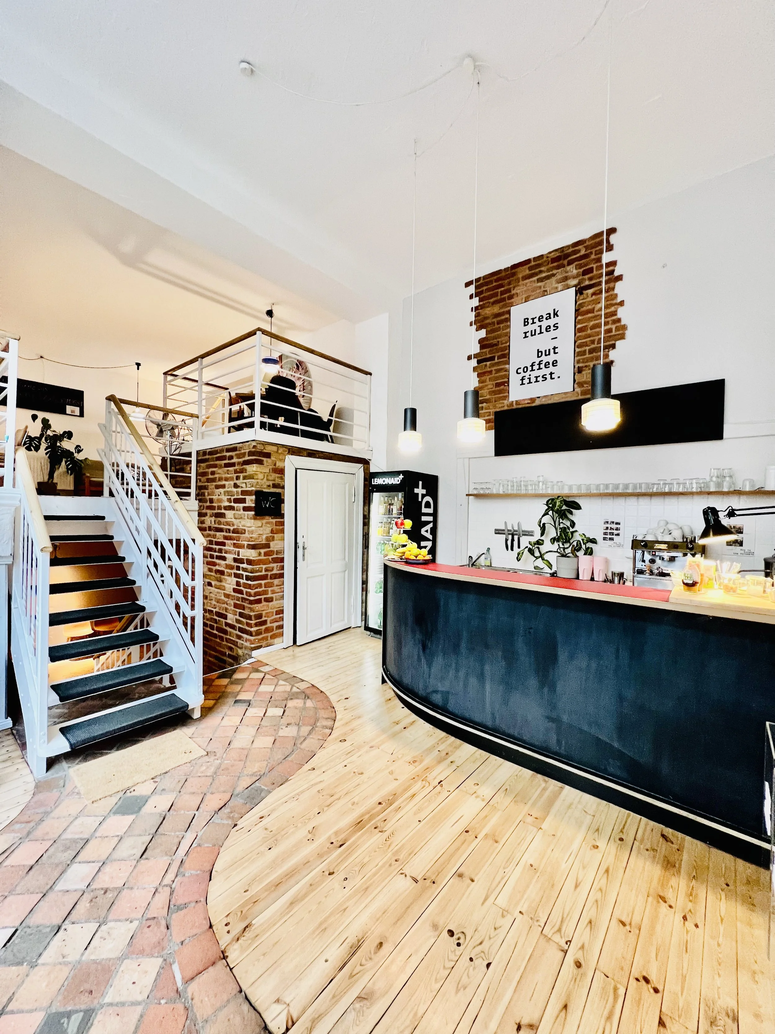 Innenbereich eines Café mit einer Theke, einem Regal mit Gläsern, einem kleinen Kühlschrank, einem Stehpult mit Früchten und einer Treppe zu einer oberen Ebene. Es gibt ein Schild, das sagt 'Breaking Rules – but coffee first'.