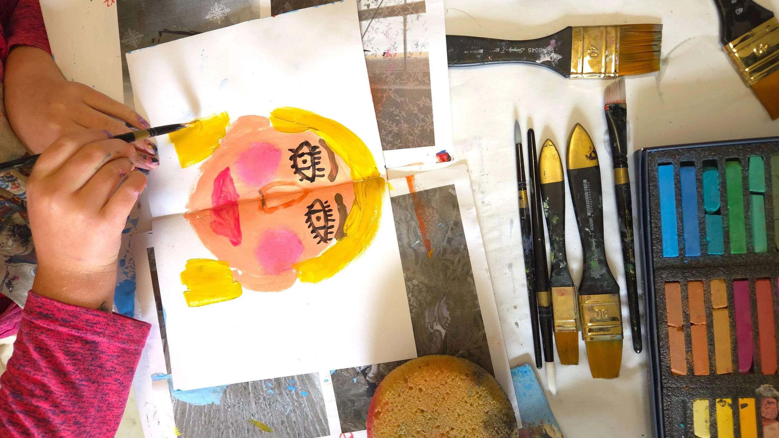 top view of little girl's hand painting a self portrait in a sketchbook, surrounded by art supplies