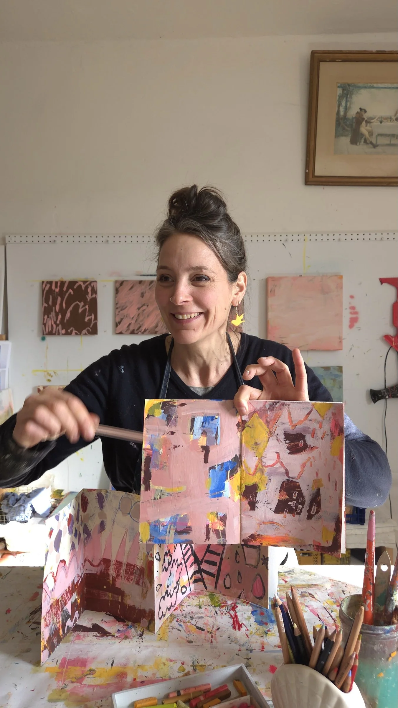 A smiling woman with dark hair in an updo, wearing a black top and yellow earrings, is working on an abstract colorful art project with pinks, blues, and blacks. She holds a paintbrush in her right hand and displays a creative, patterned, and painted artwork in a studio with paintings and art supplies around her.