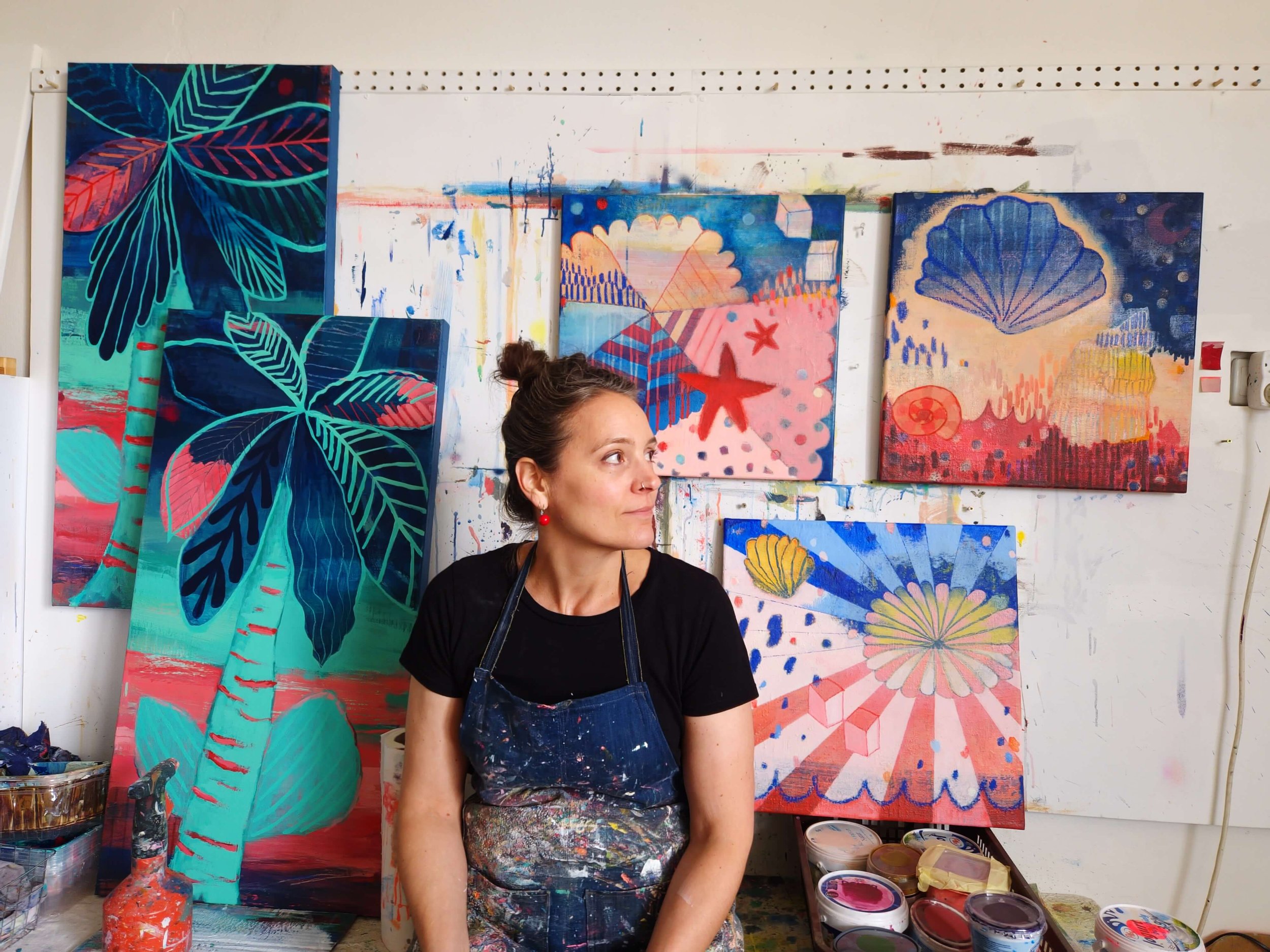 A woman in an artist's apron and black shirt stands in front of a wall with colorful abstract paintings, with art supplies like paint cans in the foreground.