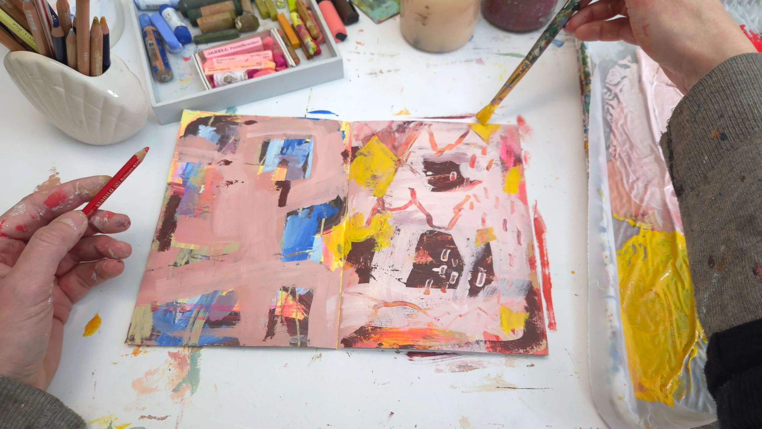 OPEN sketchbook, pages covered in paint, with the hands of the artist holding brushes