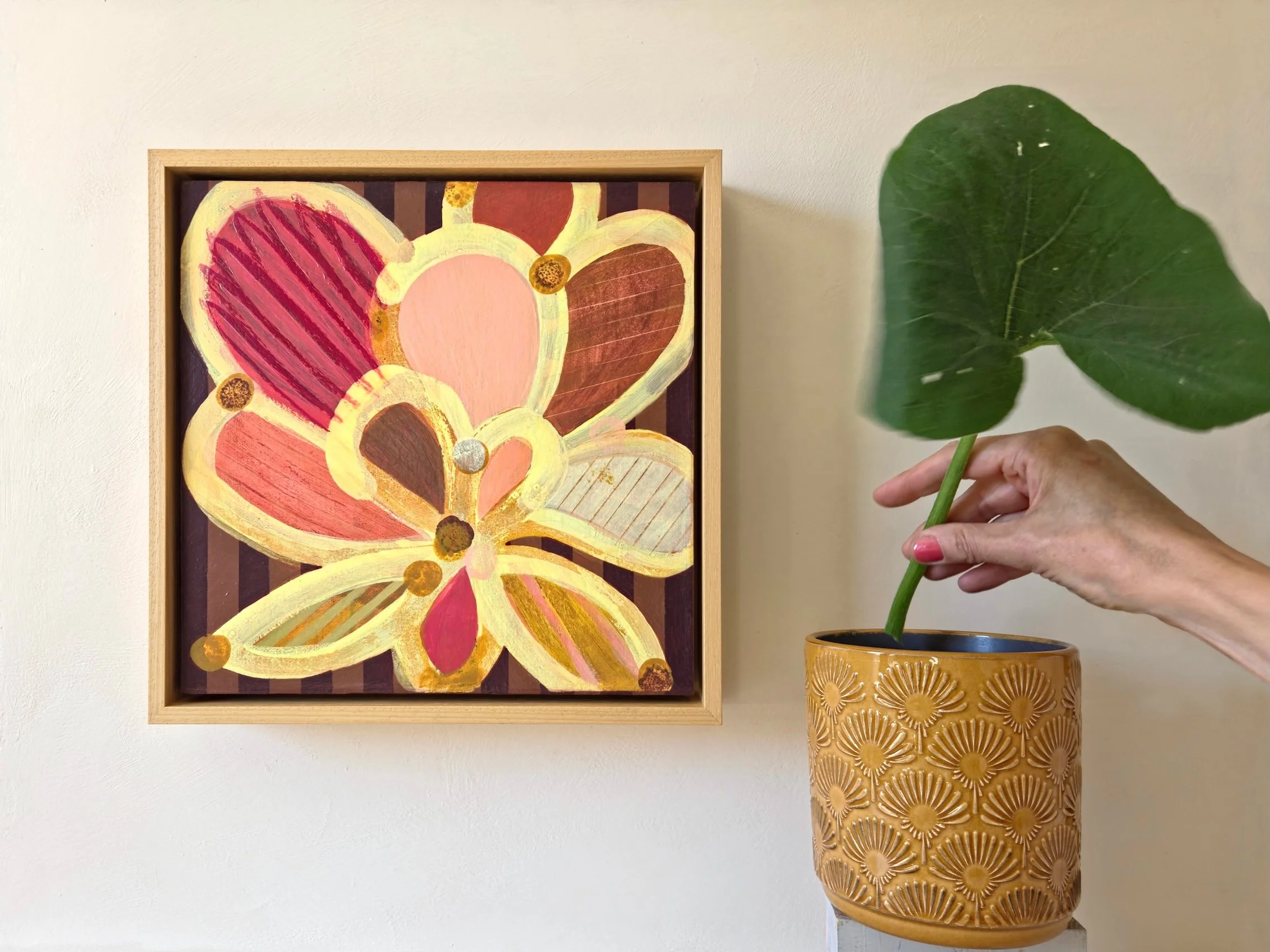 framed botanical painting in yellows and pinks on a striped background, hanged on a wall next to a vase and big leaf