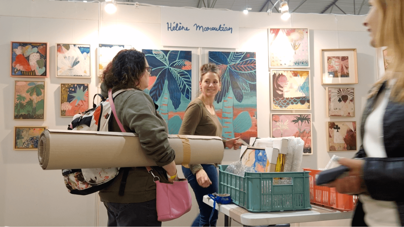 Two women are conversing at an art booth displaying colorful paintings of tropical leaves and abstract designs. One woman has curly hair, glasses, a green jacket, and is carrying a rolled-up mat and a pink bag. The other woman has straight hair, a brown shirt, and is smiling while holding a small item. A third woman on the right, partially visible, is wearing a black jacket and holds a phone or small device. There are plastic bins with bubble wrap and other items on the table in front of them. A sign with