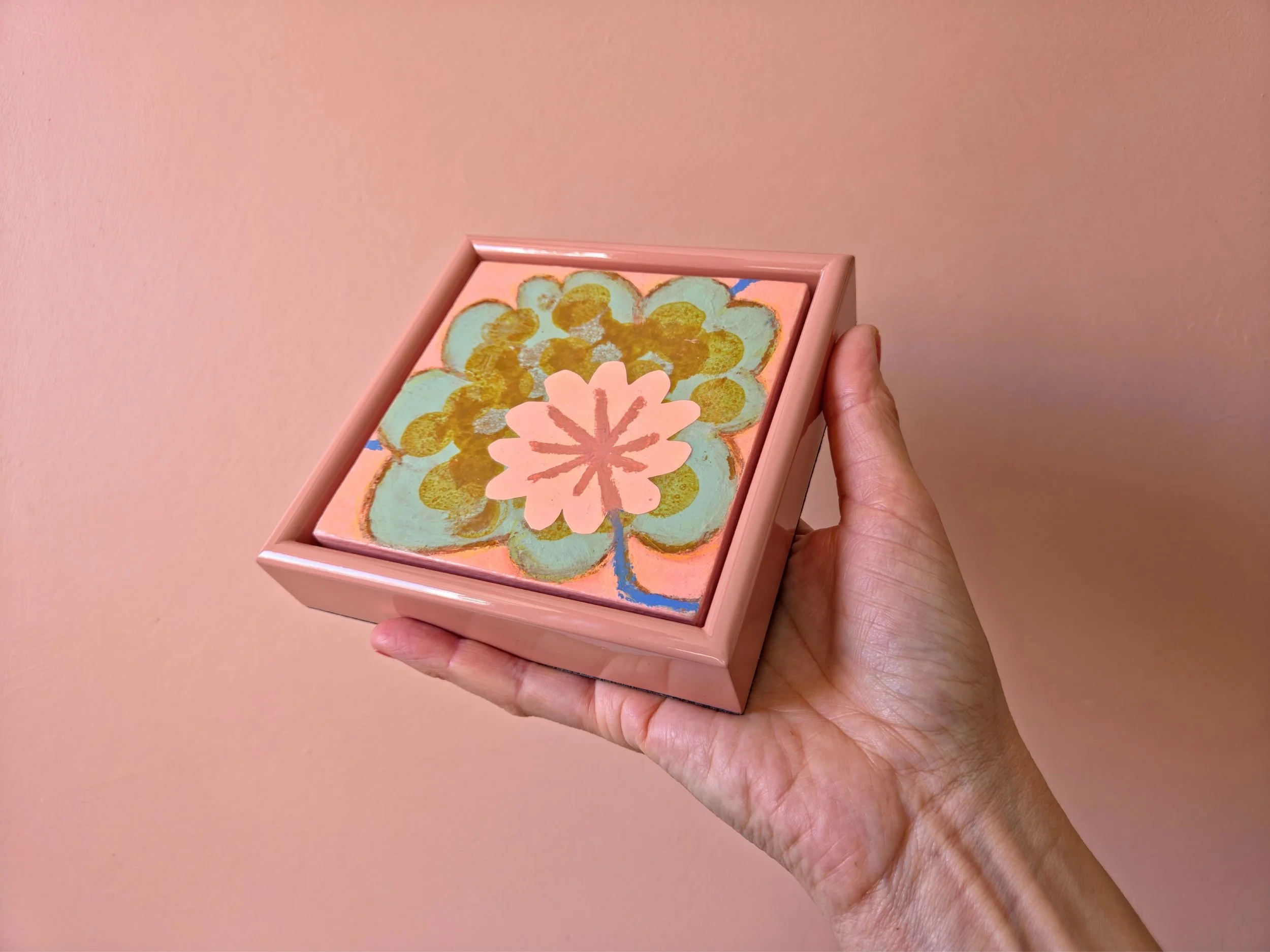 small framed botanical painting held in a hand on a pink background