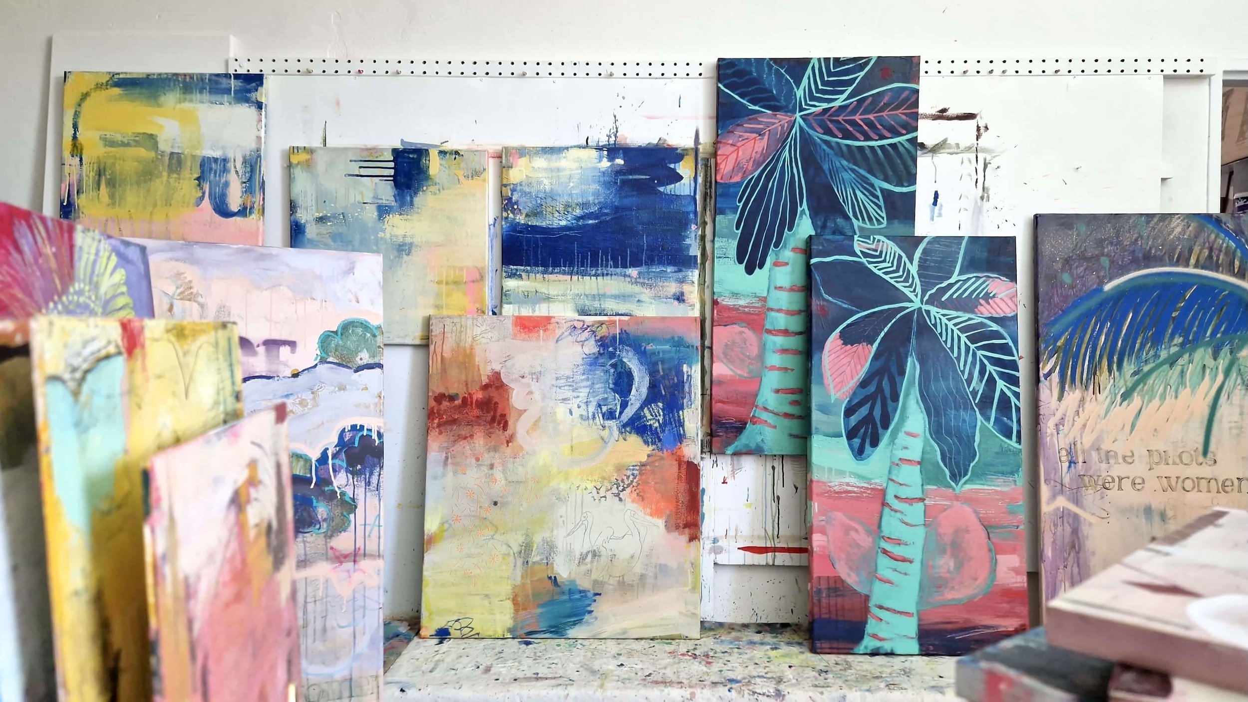 Collection of abstract and floral paintings on small canvases, arranged on a white pegboard wall, featuring vibrant colors like blue, pink, yellow, and green.