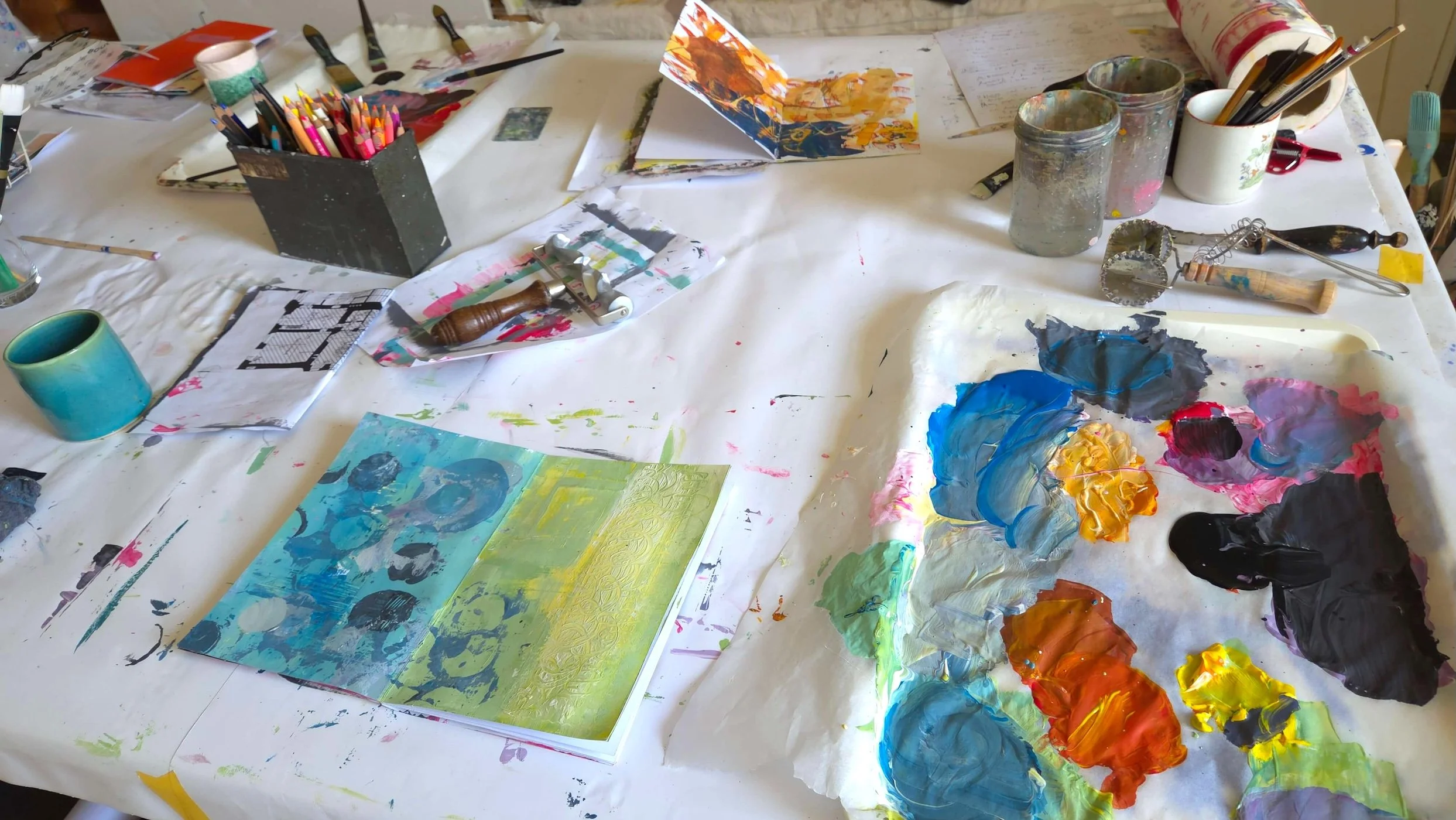 Open sketchbook on a table with painting palette and coloured pencils and more art supplies