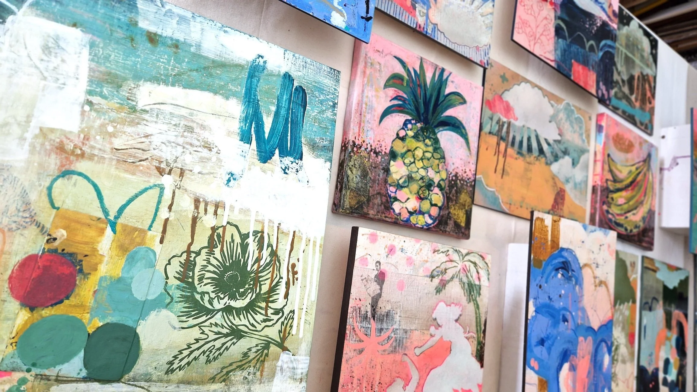 Table filled with colourful, abstract paintings including a pineapple, a man, and floral designs, waiting to be framed on the studio table