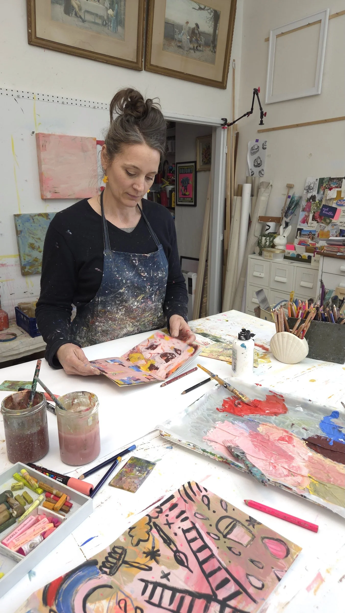 Artist with a bun hairstyle, wearing a black shirt and a paint-splattered apron, looks down at a colorful abstract art piece on her workspace. The studio has paintings on the wall, art supplies, brushes, and jars of paint on the table.
