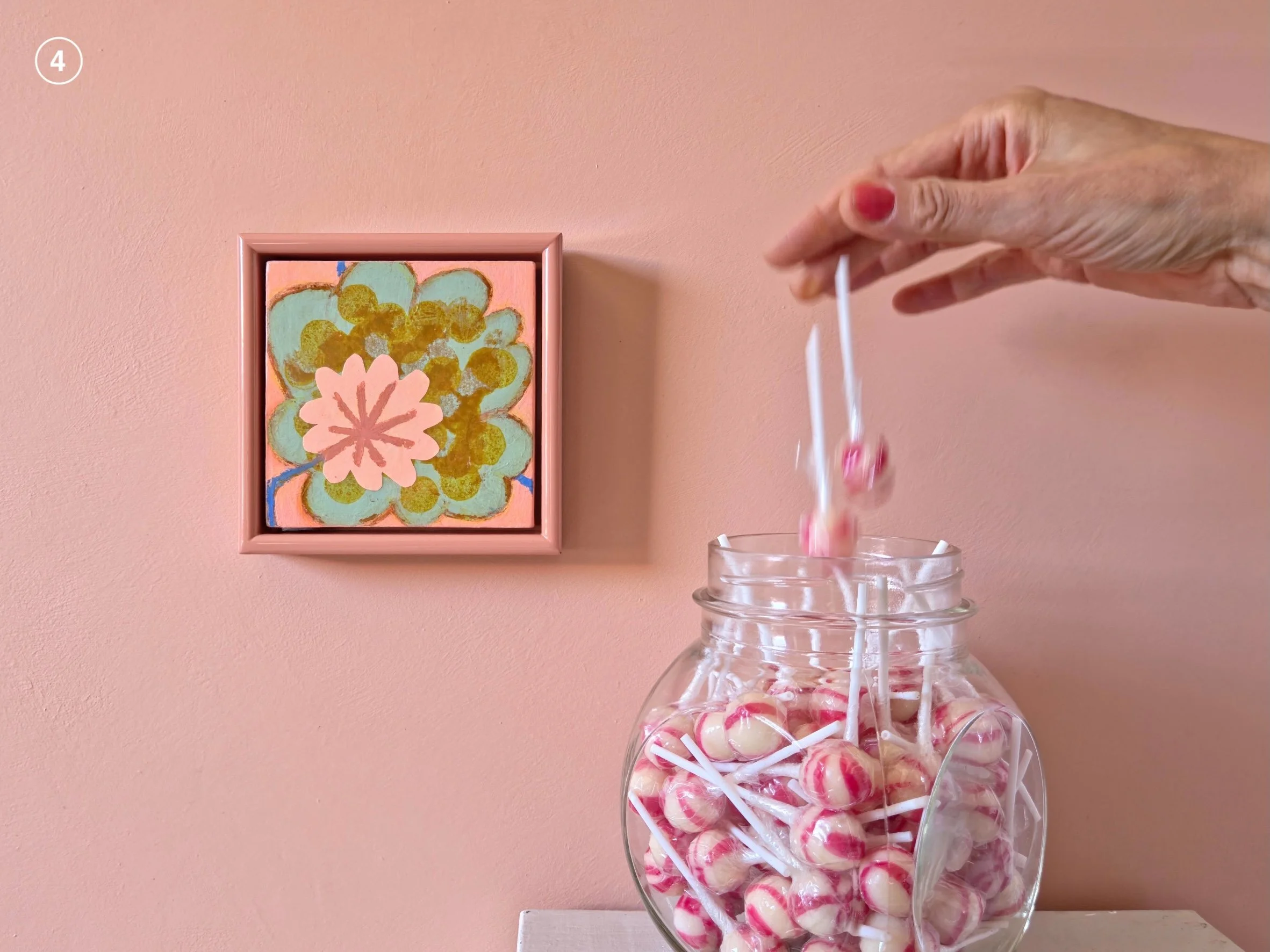 small framed floral painting on a pink wall next to jar of candies
