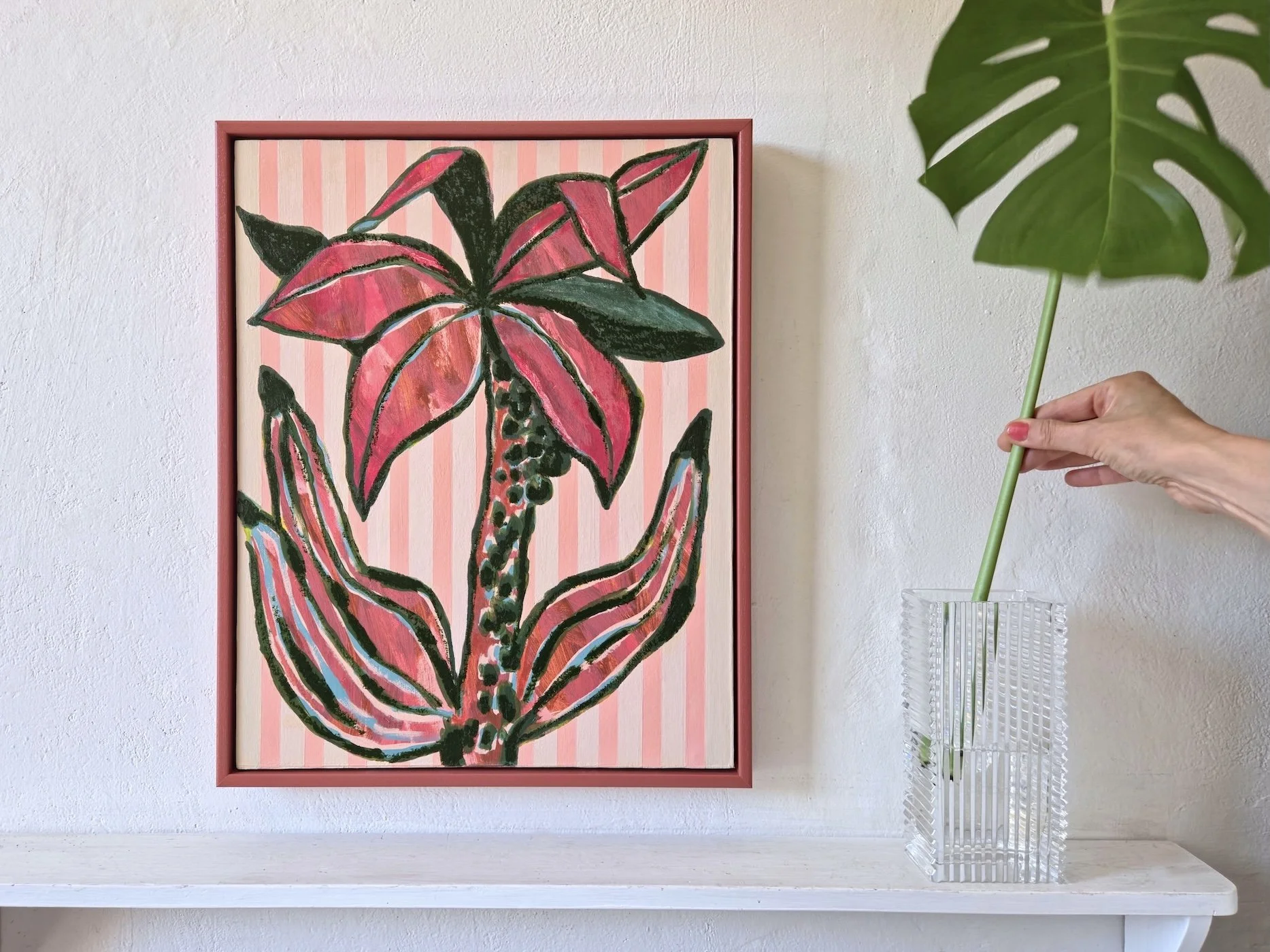 framed palm tree painting with pink stripes background with a vase and monstera leaf