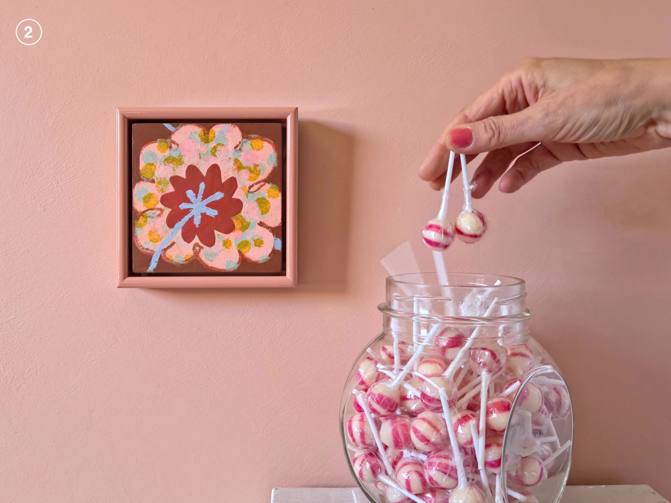 small framed painting of a flower on a pink wall next to jar of candies
