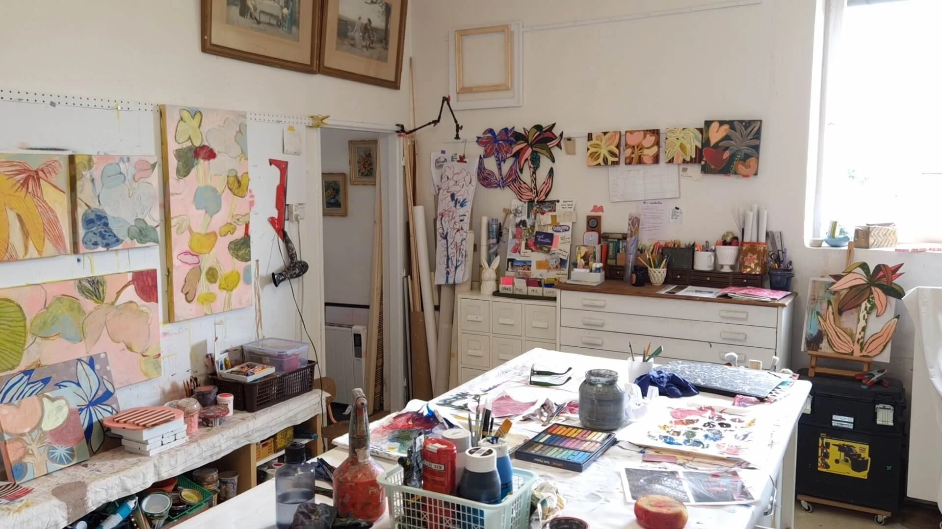 wide shot of an artist studio, with a big messy table n the middle, covered with material and walls covered with canvases in progress