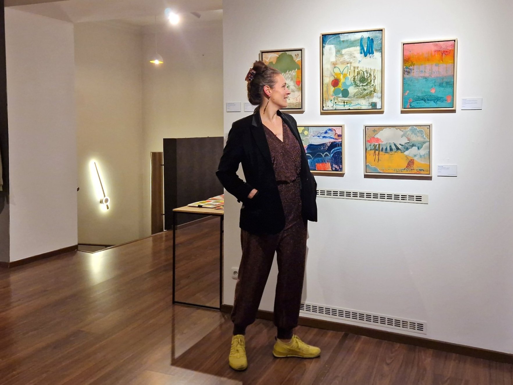 Woman in black blazer and glitter pants standing in an art gallery, looking at colourful abstract paintings on the wall. She is wearing bright yellow shoes and has her hair up in a bun.