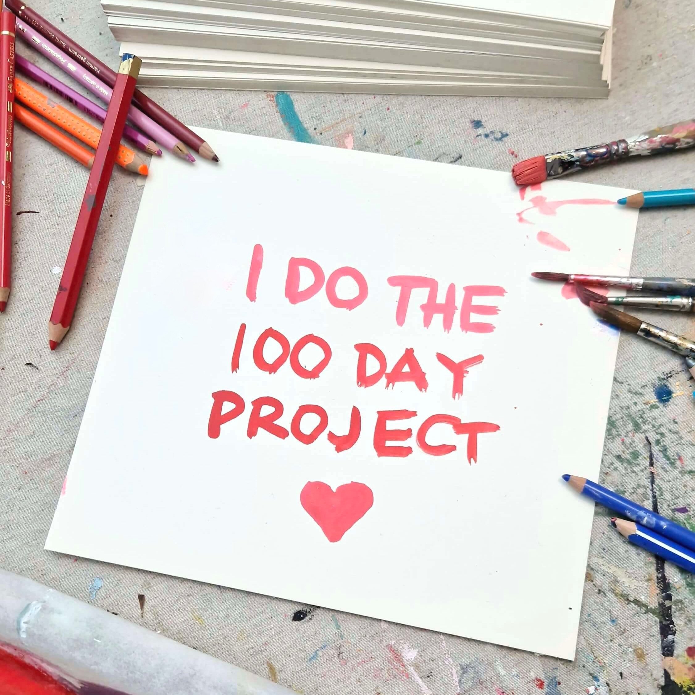 Studio table with a text painted in red on a paper I do the 100 day project, with brishes and pencils