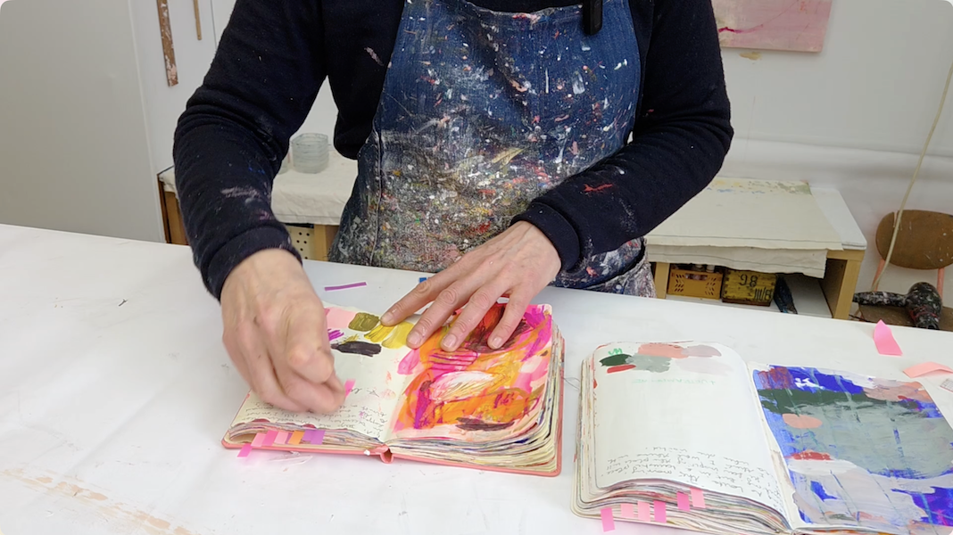 an artist selecting pages in two sketchbooks on a table in the studio