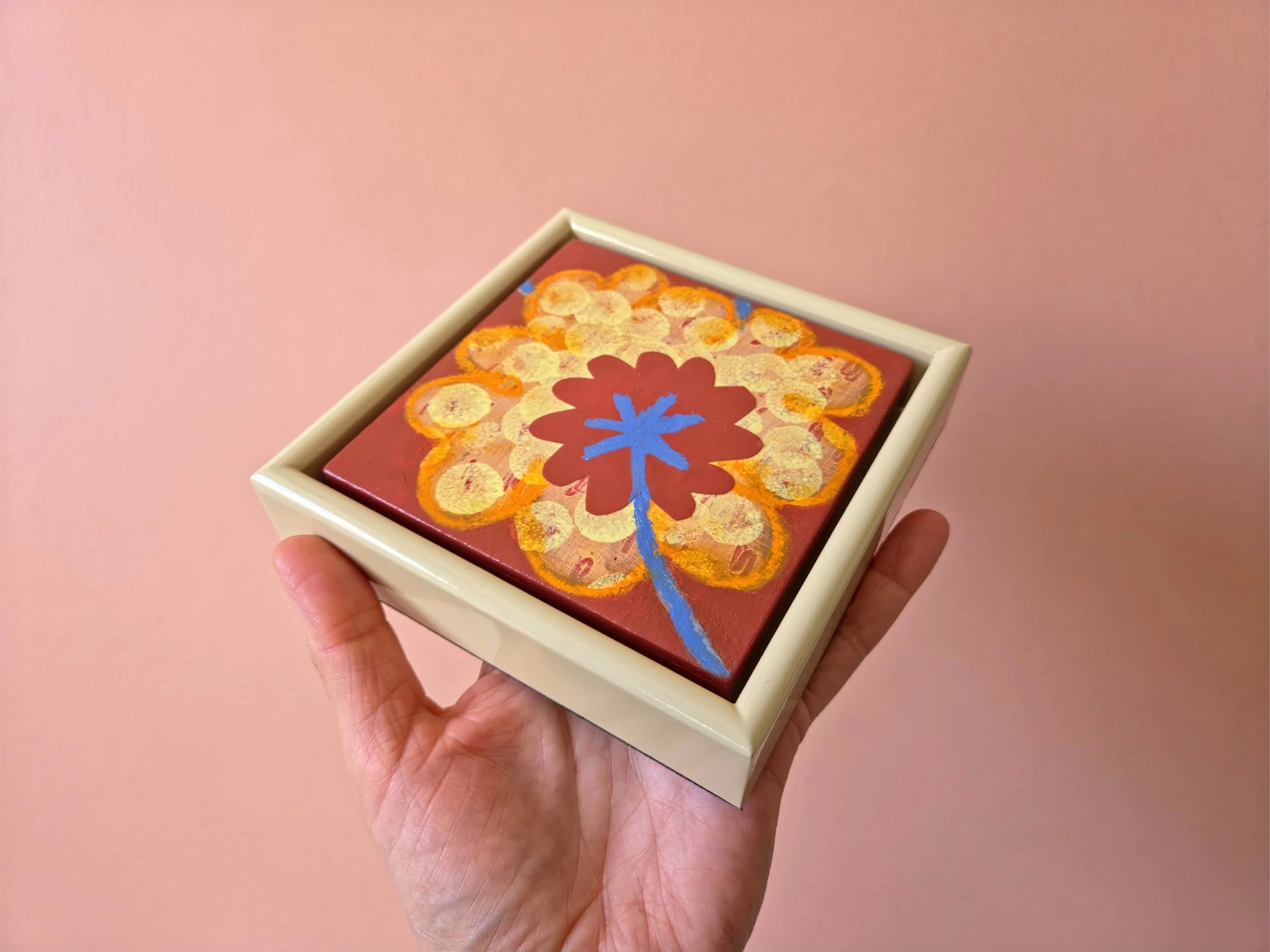 small framed botanical painting held in a hand on a pink background