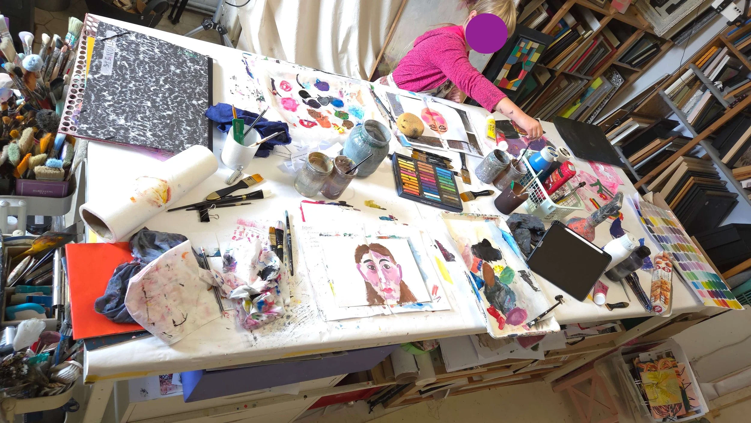 Big table in an artist studio with workshop in progress, on participant, a child and a lot or art supplies