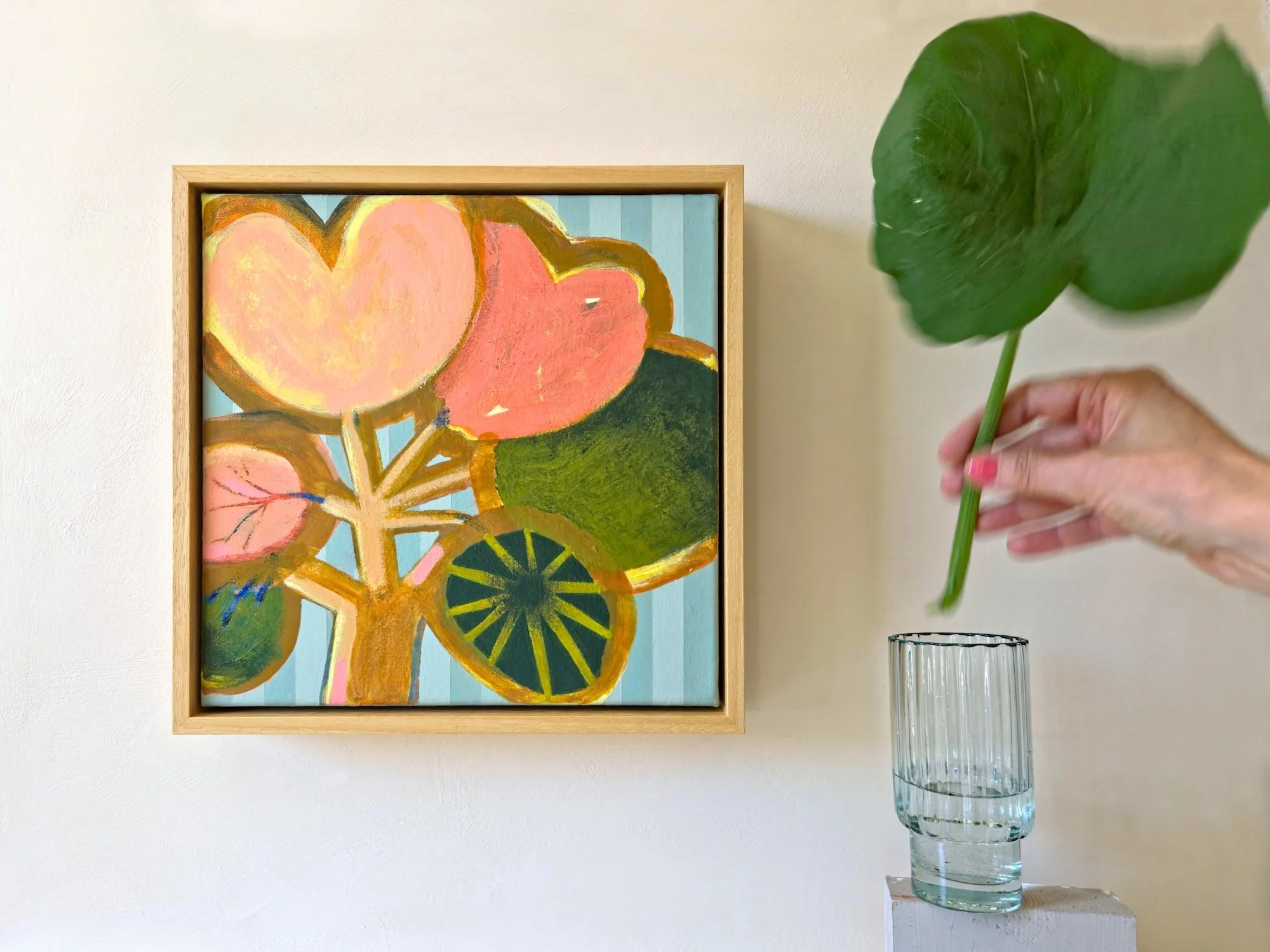 framed botanical painting in bright pinks and greens on a striped background hanged on a wall next to a vase and big leaf