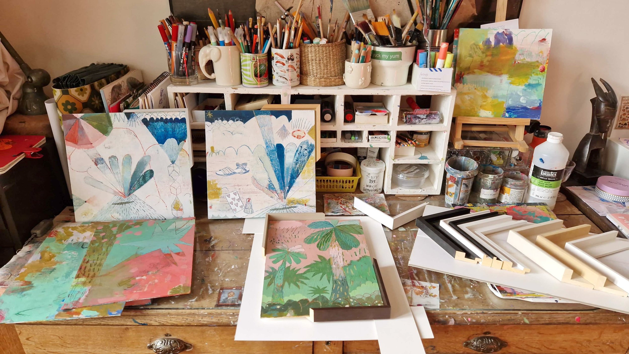 An art workspace with paintings, brushes, and art supplies. Painted canvases with colorful tropical and abstract designs are displayed on a table and leaning against a white organizer filled with art tools, paints, and jars.