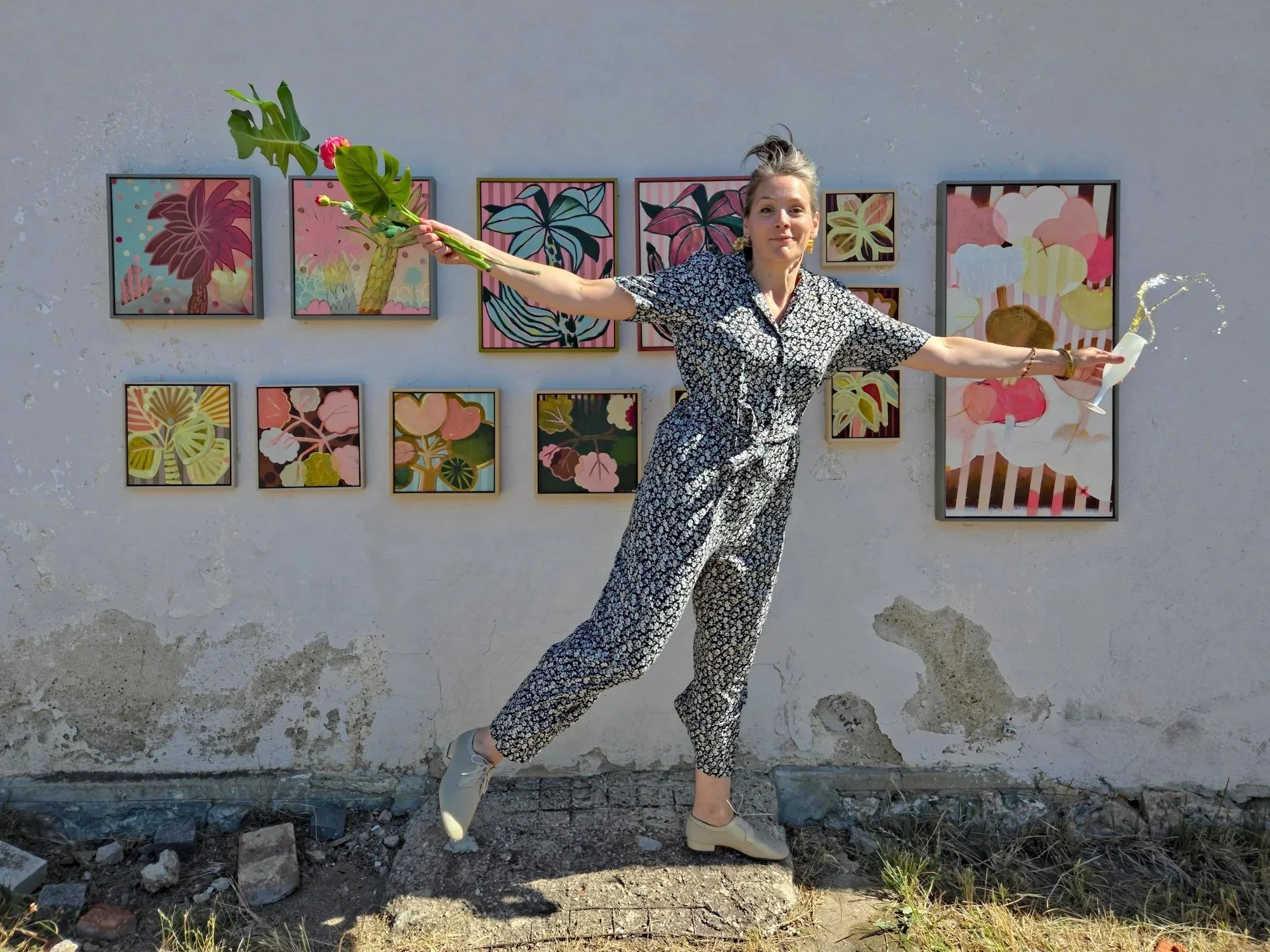 woman standng in front of a wall full of colourful botanical paintings and holding a glass of champagne in one hand and flowers in the other