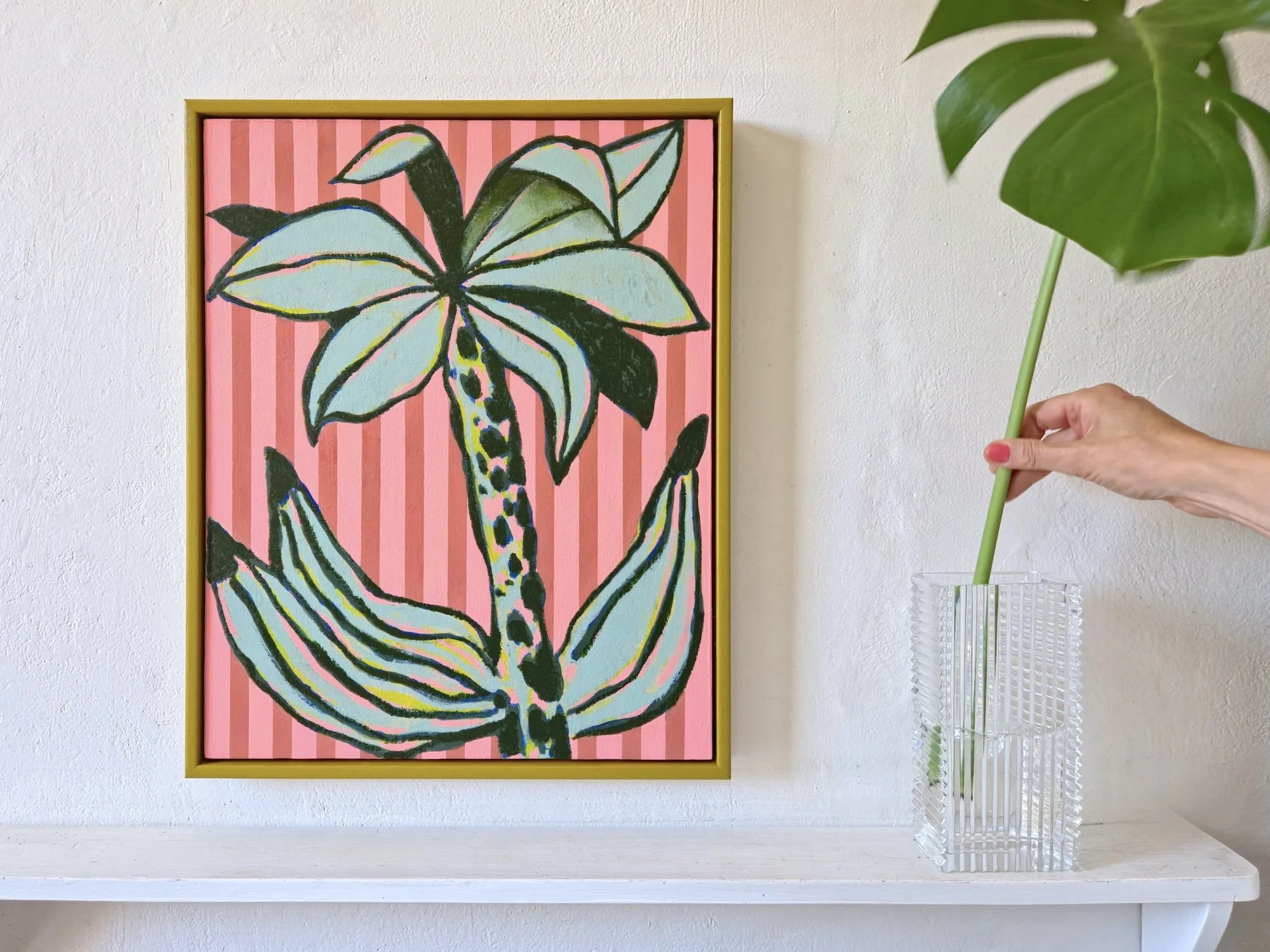 framed palm tree painting with pink striped background and a vase with monstera leaf