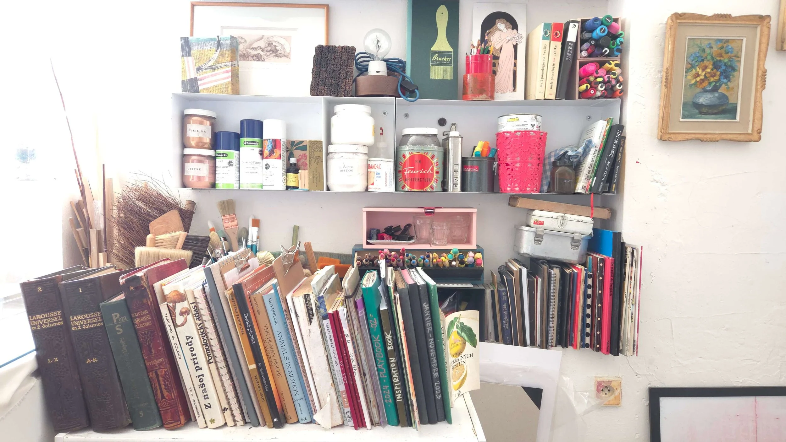 messy artist studio corner with books on a chelf and bruches in pots and all sorts of sprays