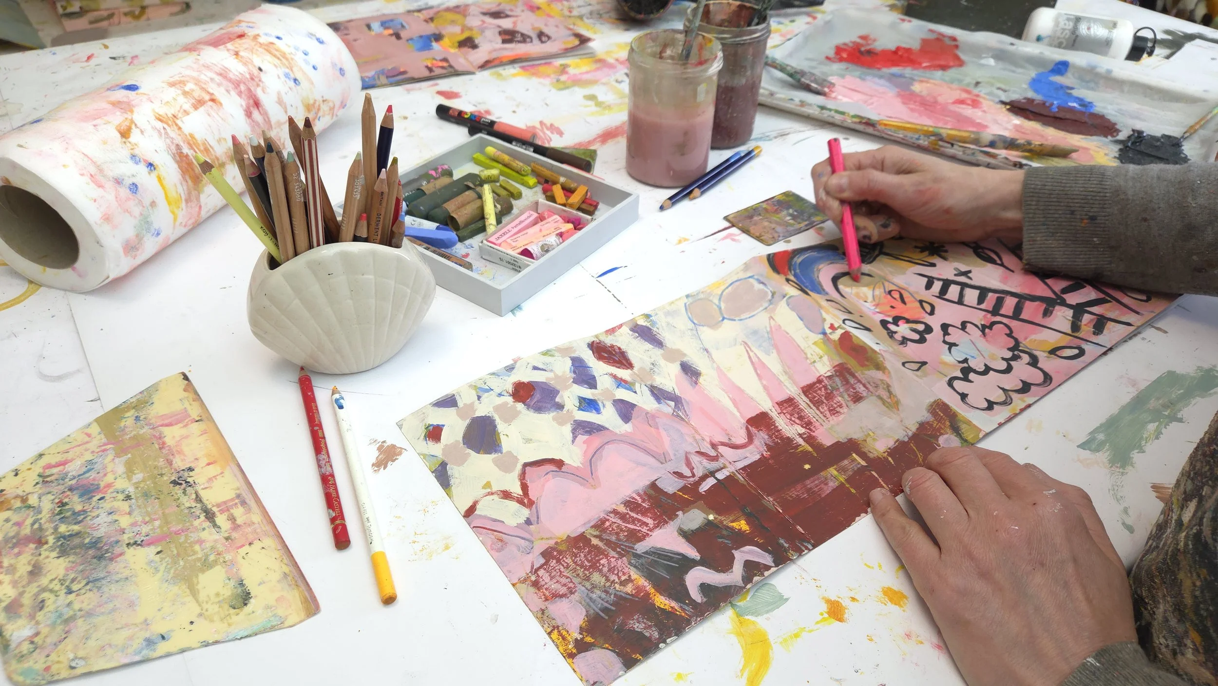 A person creating abstract paintings on paper with colorful paints, pencils, and brushes on a cluttered table.