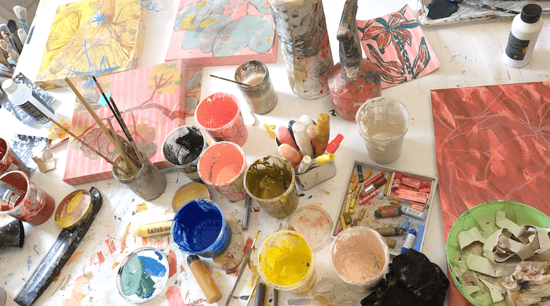 a table covered with paint tubs, colourful pastels, brushes and art making tools