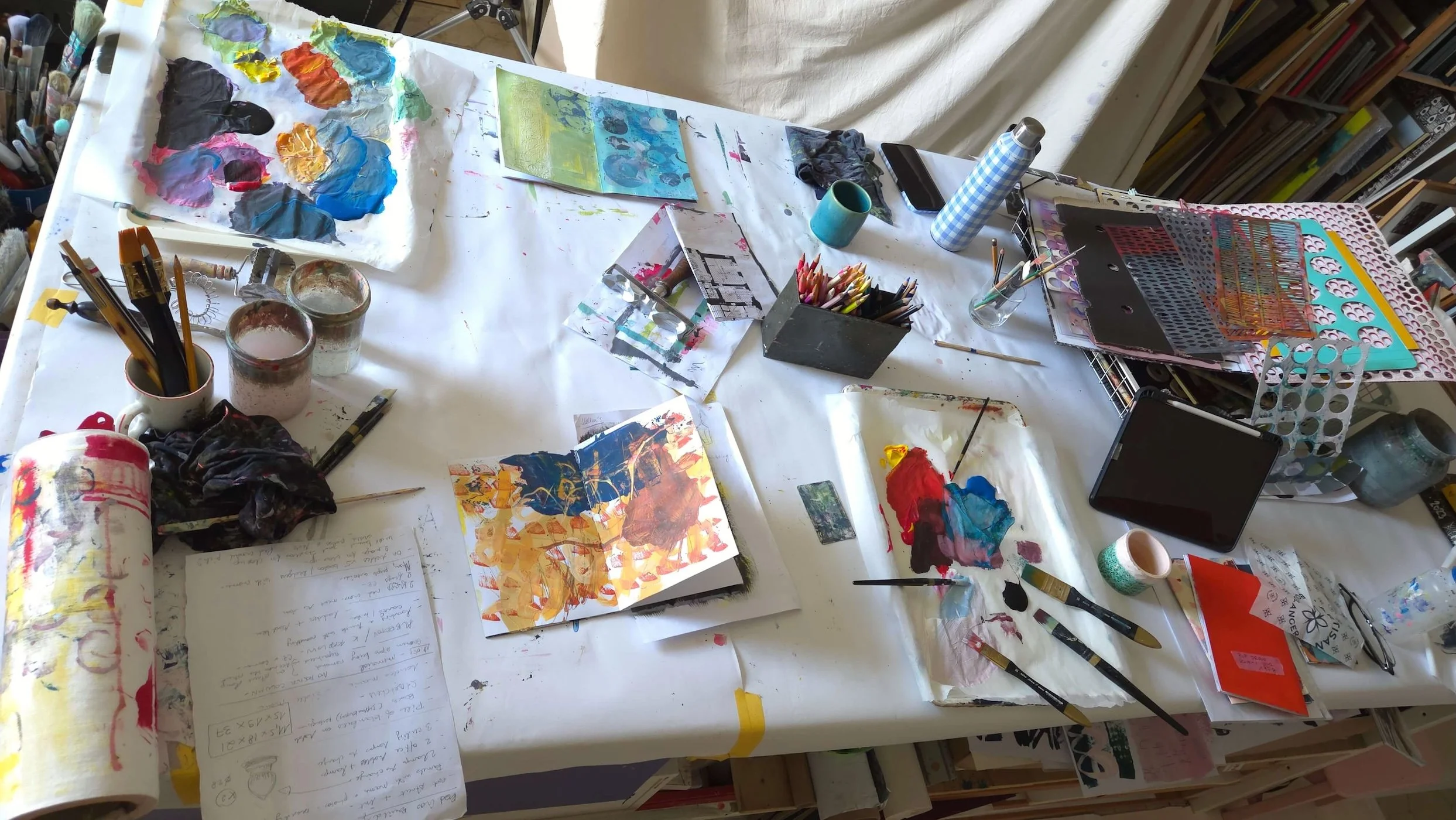 Big table with open sketchbooks, paint palettes, coloured pencils and other art supplies