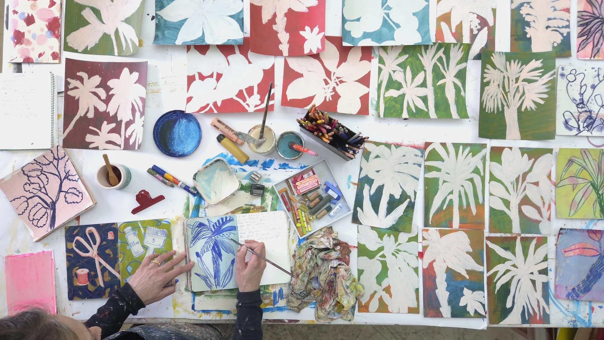 a table full of studies on paper, showing various versions of palm trees in colour with tools and paintbrushes and the artist's hands