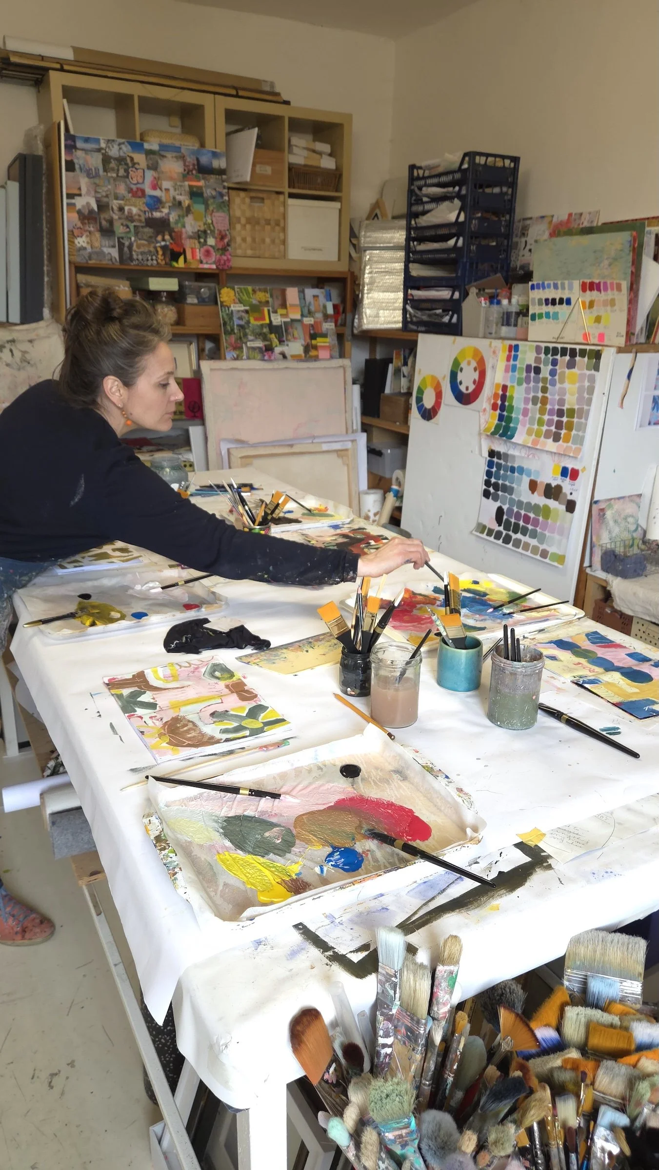 An artist working at a table covered with paint palettes, brushes, and paintings in progress. The background has shelves with art supplies, color charts, and completed artwork.