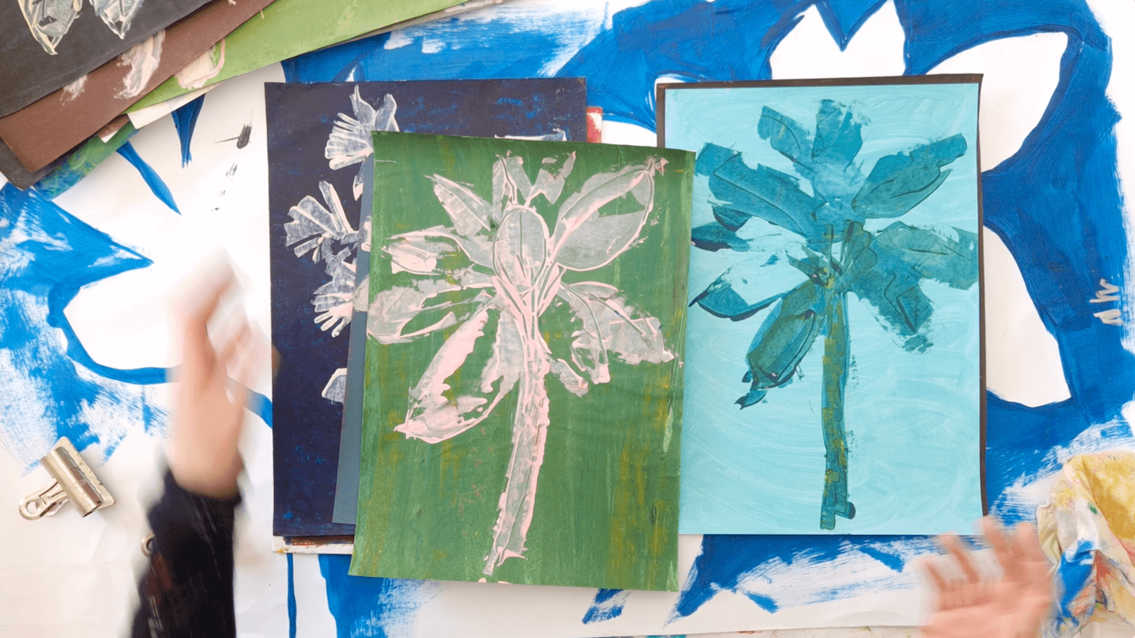 Artist sorting and reviewing colour studies of palm trees on a table, selecting favourite pieces before developing a colour palette for a complete series.