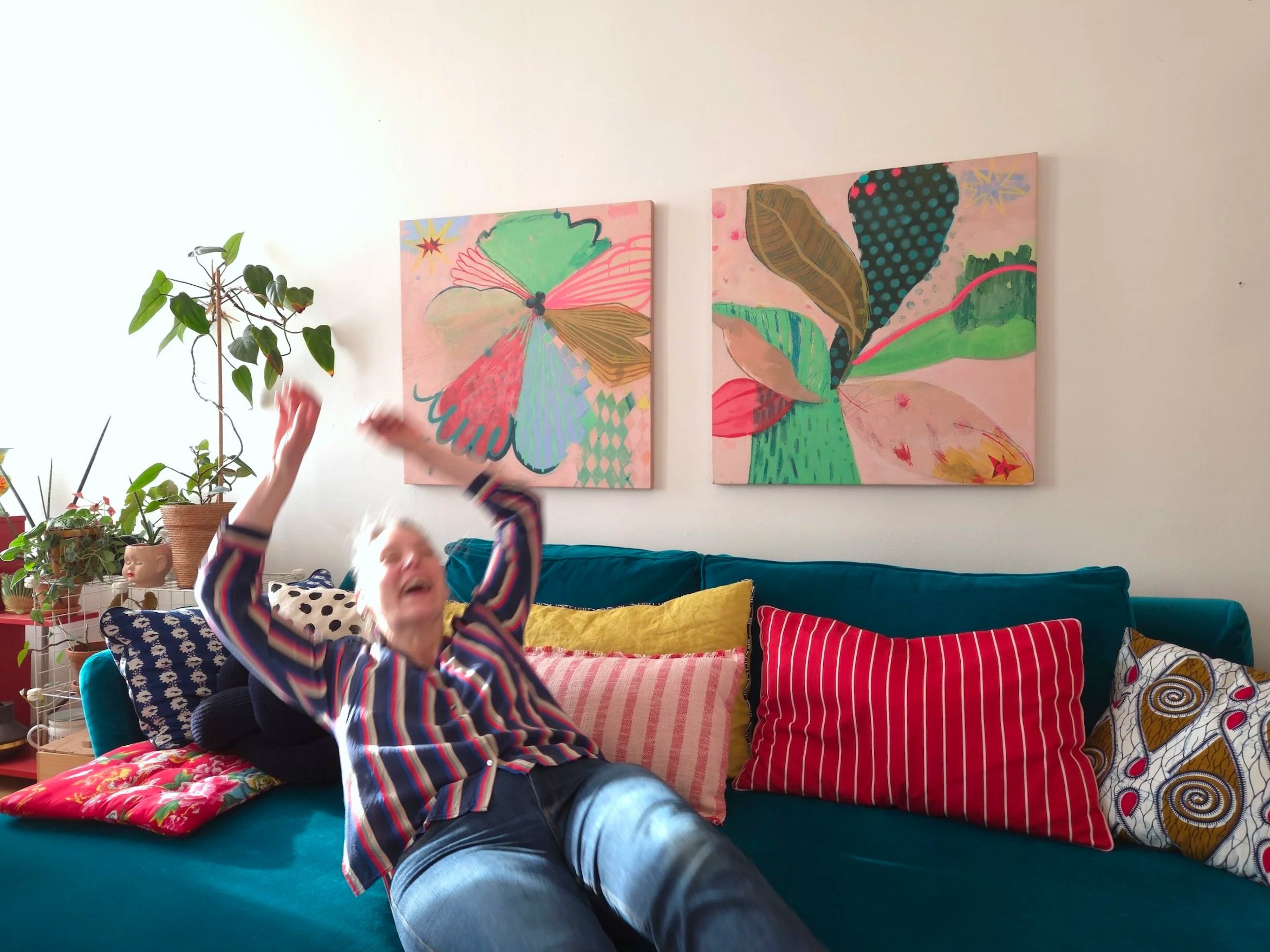 Woman wearing a striped long-sleeve shirt, relaxed on a blue couch in a brightly decorated living room, with colorful cushions, artwork on the wall, and potted plants nearby.