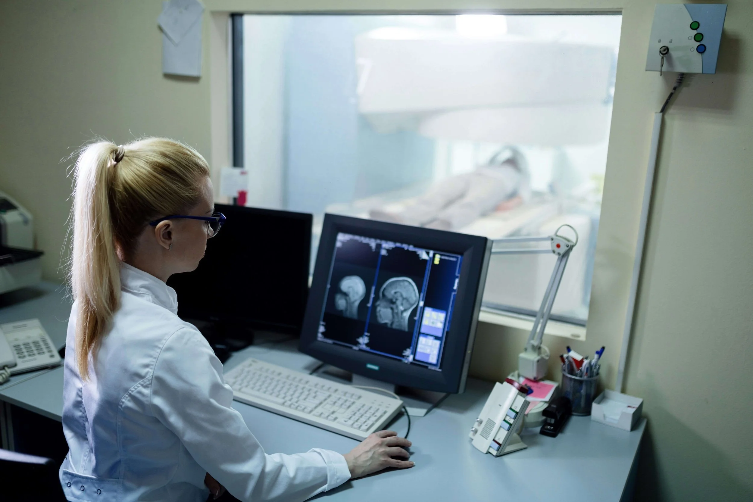 Medical professional in a white coat working on brain scan images on a computer in a hospital or clinic setting, with a patient lying down on a bed visible through a window in the background.