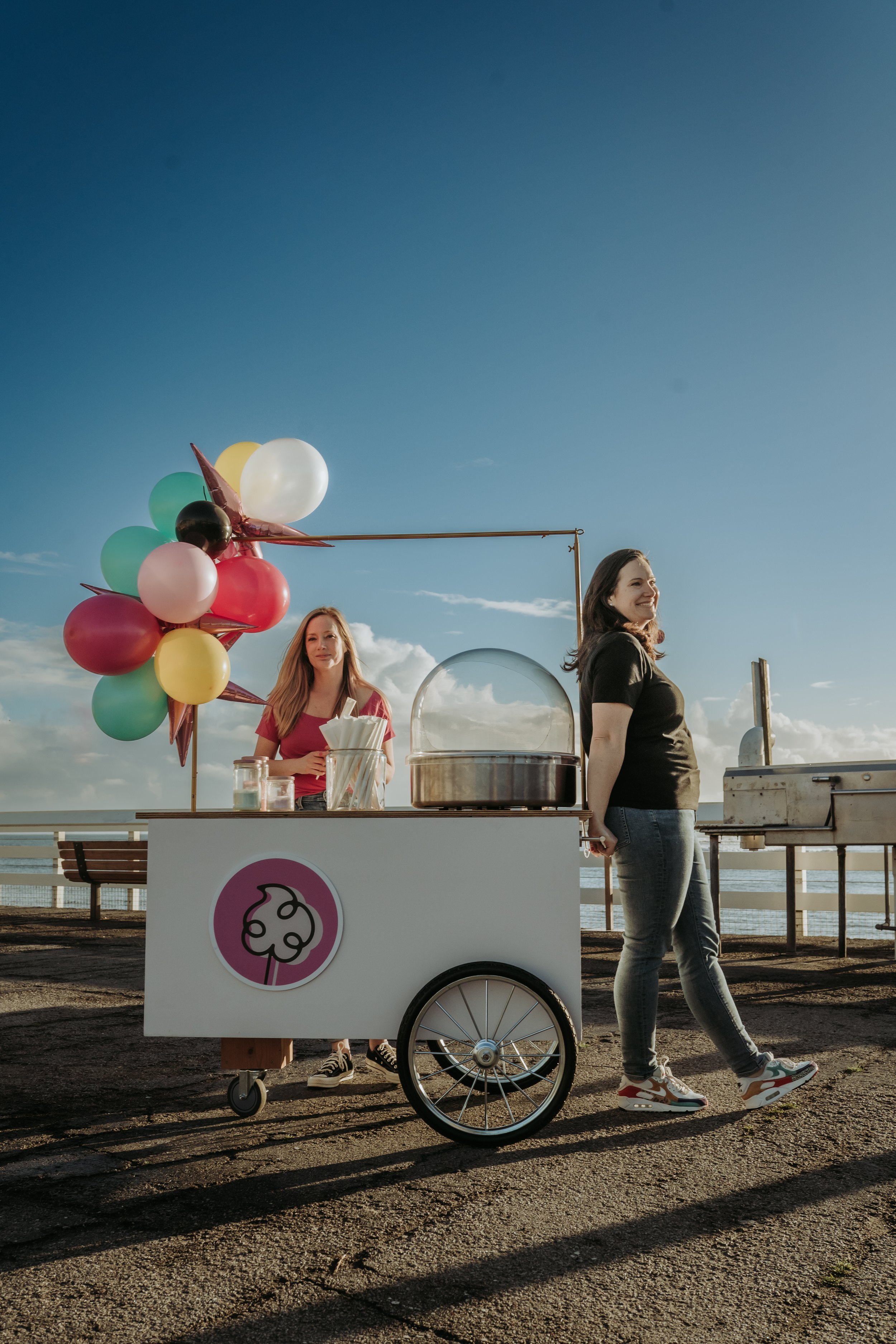 A woman standing beside a cotton candy cart on a beach, with a woman behind the cart, and balloons attached to the cart, under a blue sky with some clouds.