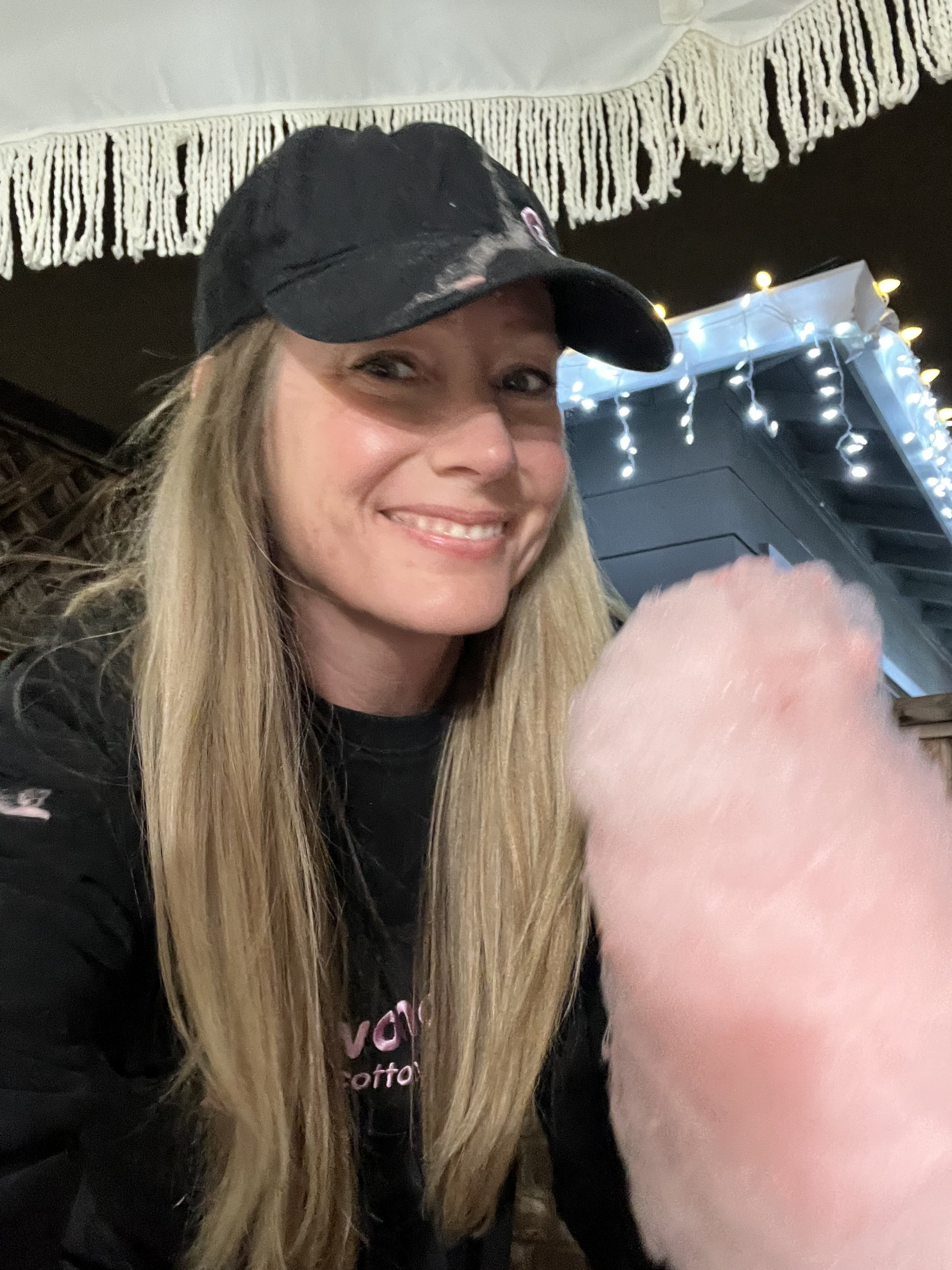 A smiling woman with long blonde hair wearing a black baseball cap and black shirt, sitting indoors near a pink cotton candy.
