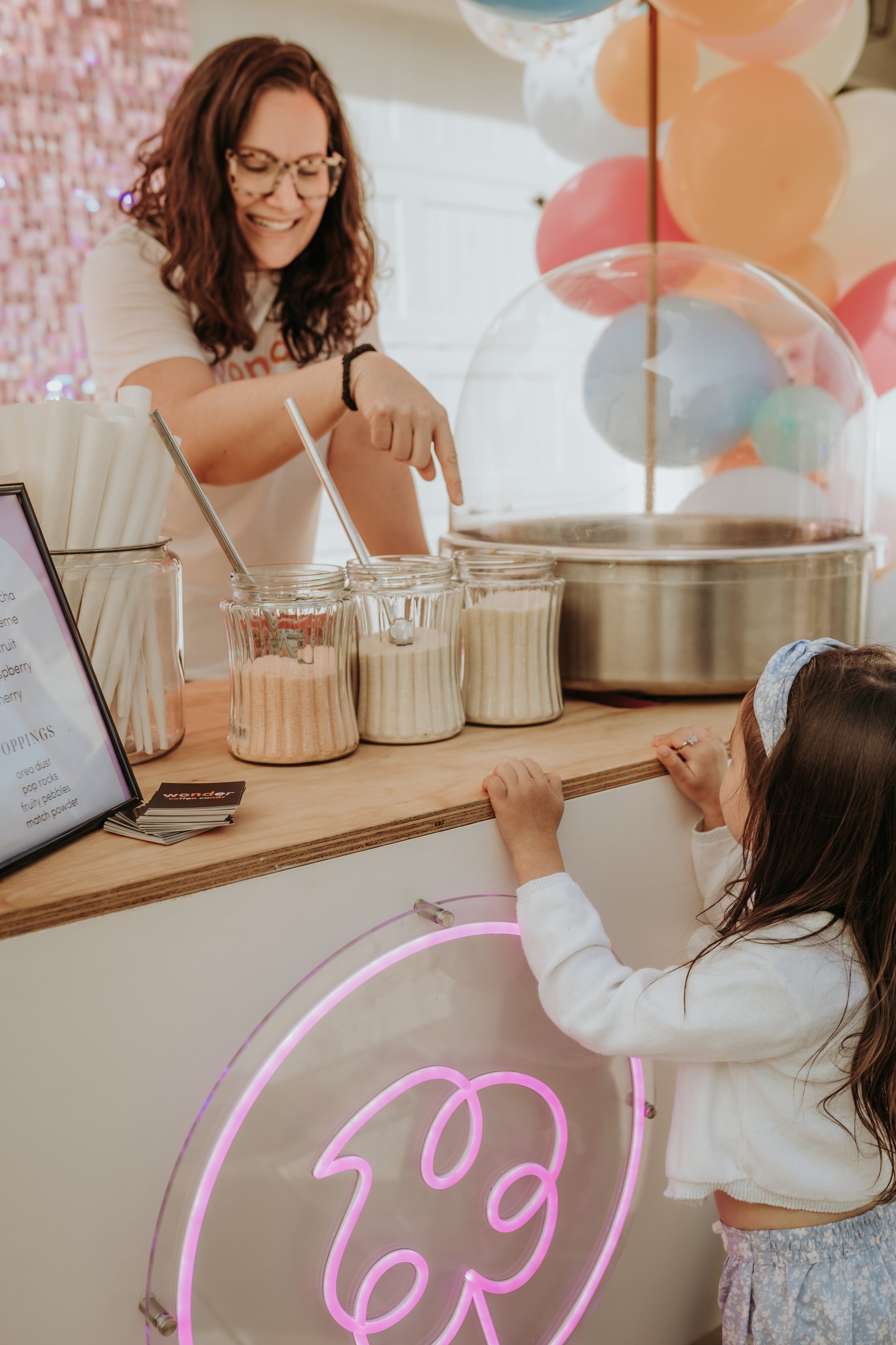 A woman with curly hair, glasses, and a white T-shirt is smiling and pointing at jars of cotton candy at a cotton candy stand. A young girl with long brown hair, a blue headband, and a white sweater is looking up at her. The stand is decorated with p