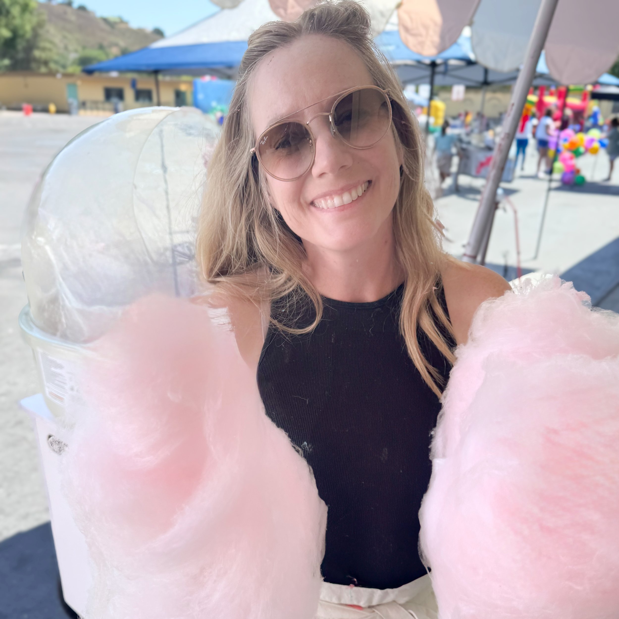Smiling woman wearing sunglasses at an outdoor fair holding pink cotton candy.