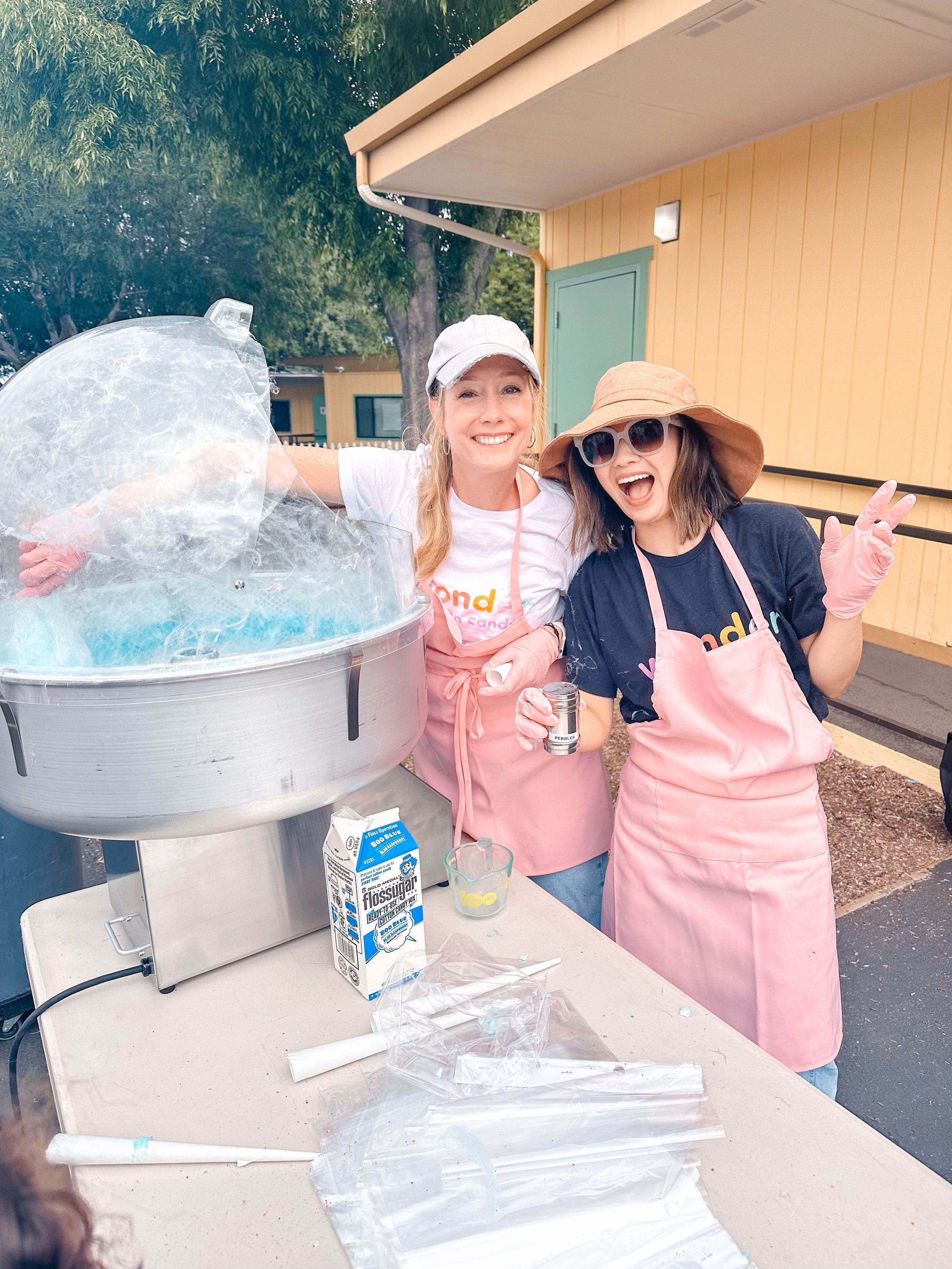 Two women smiling and posing next to a cotton candy machine at an outdoor event, wearing aprons, gloves, hats, and sunglasses, with colorful cotton candy in a cup on the table.