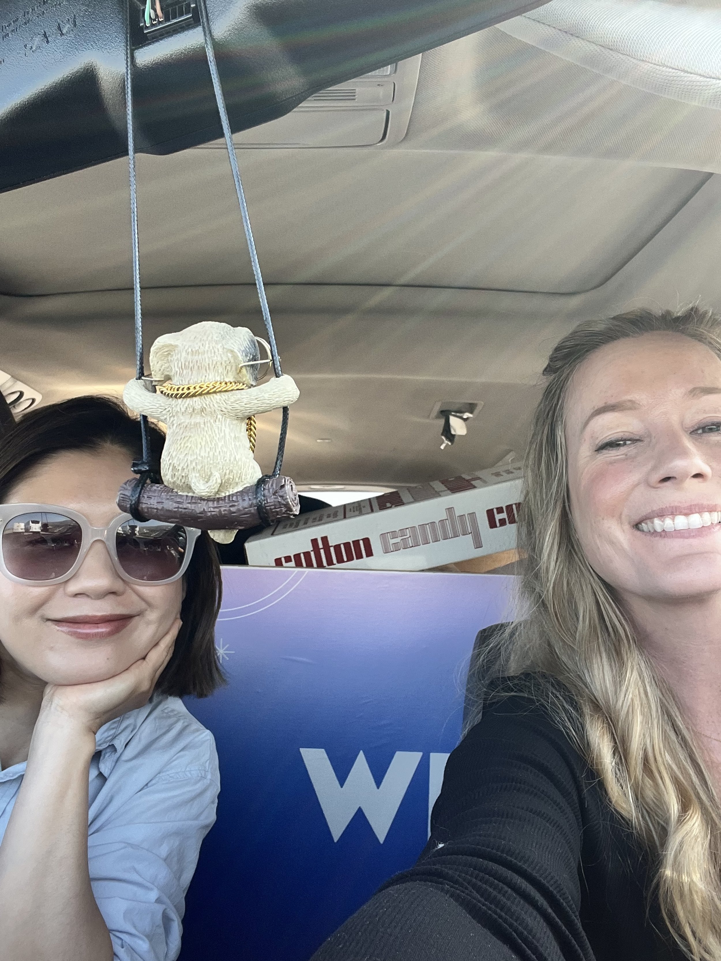 Two women sitting inside a vehicle, one wearing sunglasses and resting her chin on her hand, the other smiling. A decorative bear figurine wearing sunglasses, a hat, and a gold chain hangs from the ceiling. Behind them are boxes including one labeled
