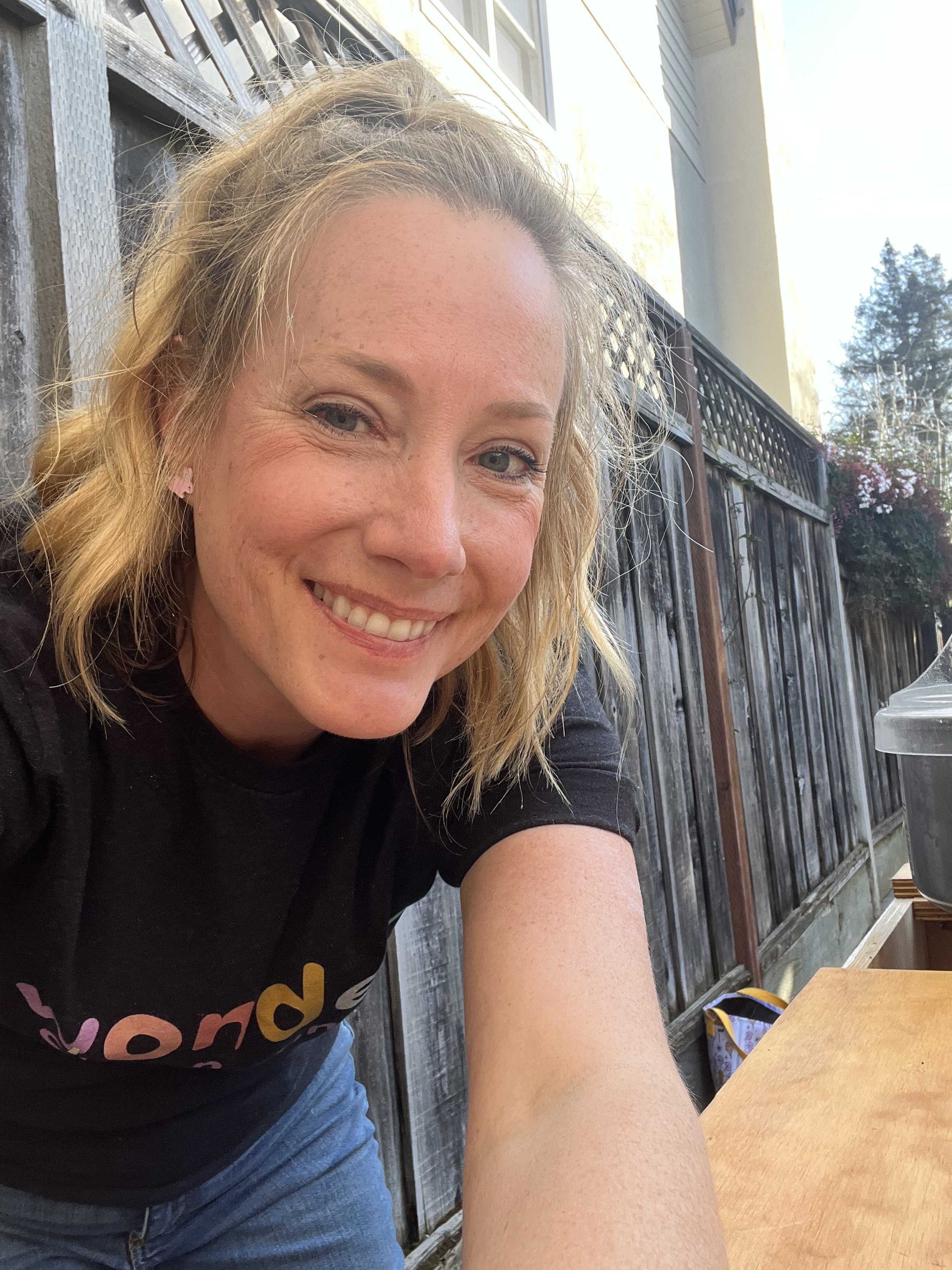 A young woman with blonde hair smiling while taking a selfie outdoors near a wooden fence and a house, with a table and a drink in front of her.
