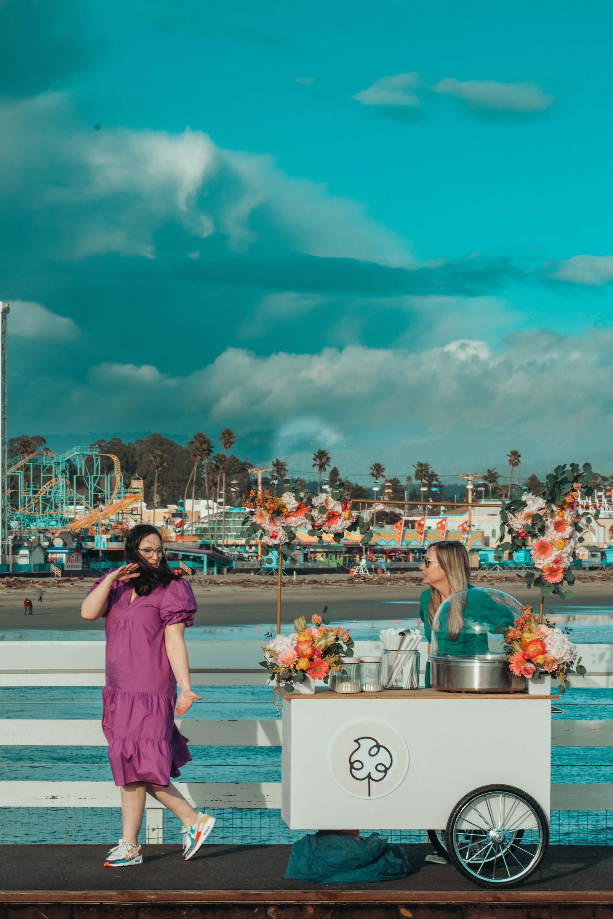 A woman in a purple dress walking past a floral ice cream cart on a boardwalk by the beach, with amusement park rides and palm trees in the background under a partly cloudy sky.