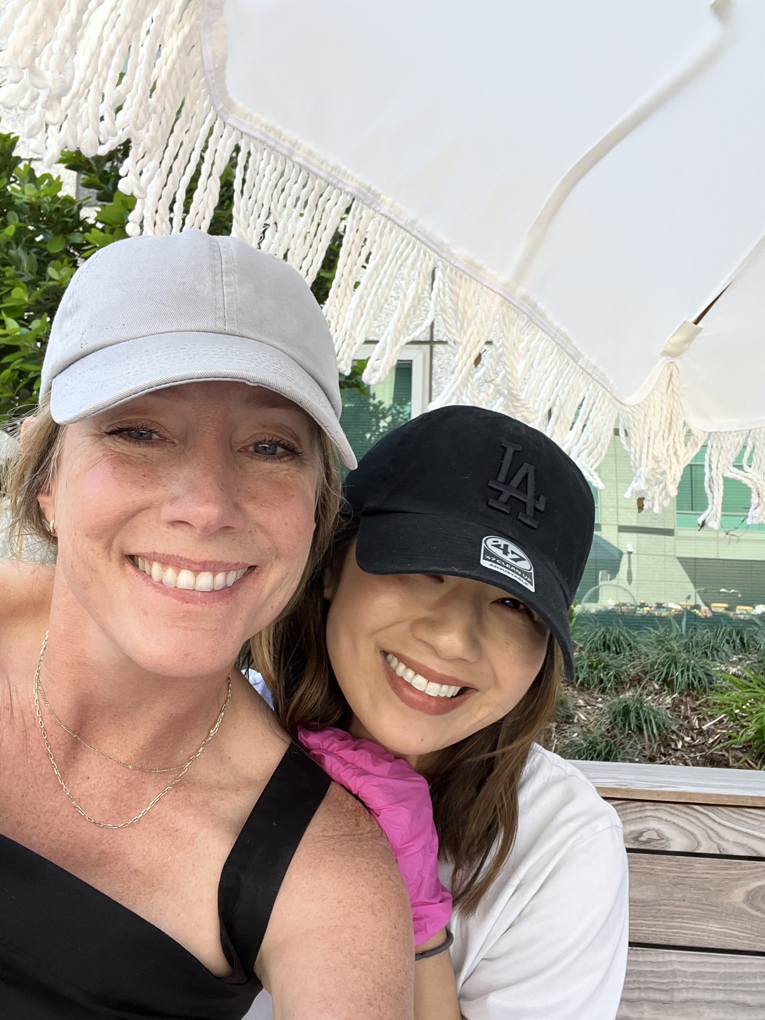 Two smiling women wearing baseball caps, sitting close together outdoors under a white umbrella.