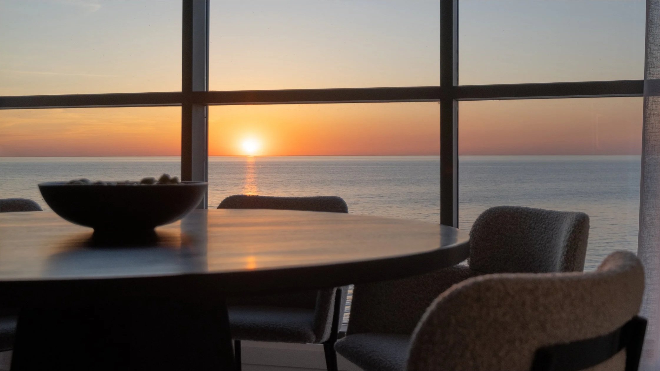 Sunset Condo. Indoor dining area with a round table and chairs, overlooking the ocean and a sunset through large windowpanes.
