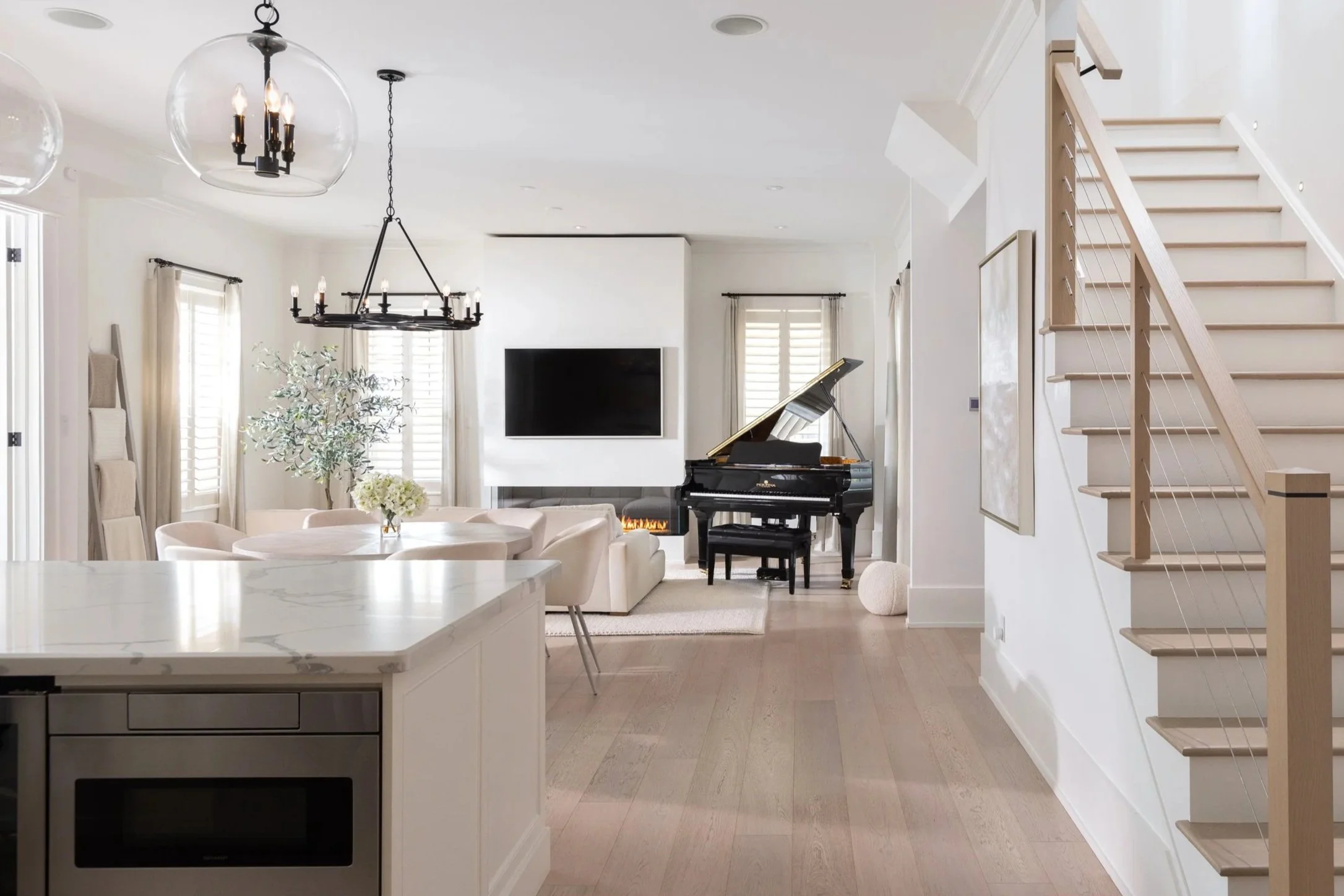 Modern living room with a grand piano, a wall-mounted TV above a fireplace, a dining table with chairs, and a staircase on the right side.