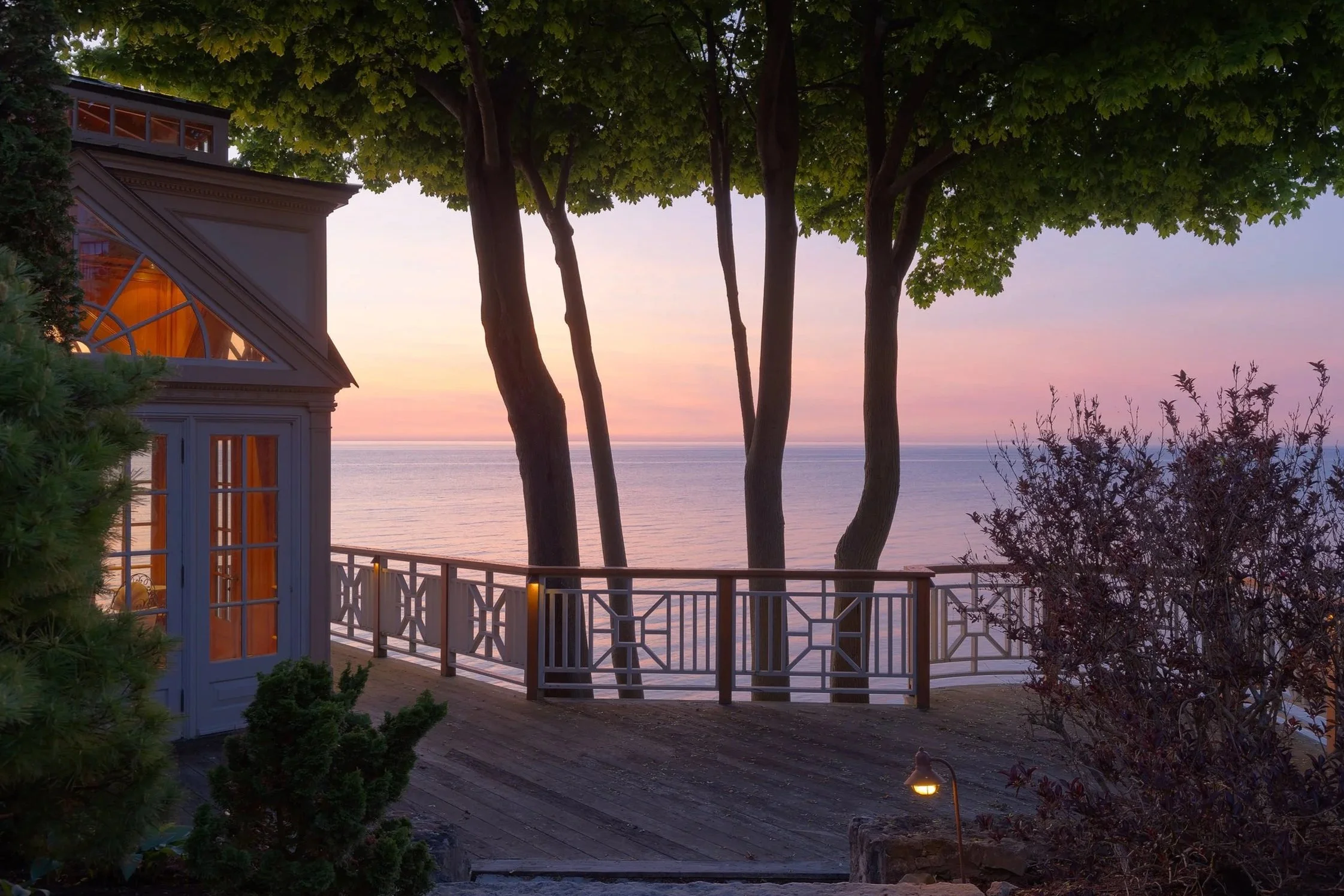 A porch or balcony with a wooden railing overlooking a calm ocean at sunset. There are trees framing the scene, and a house with large windows to the left, partially visible. The sky displays soft pink and purple hues.