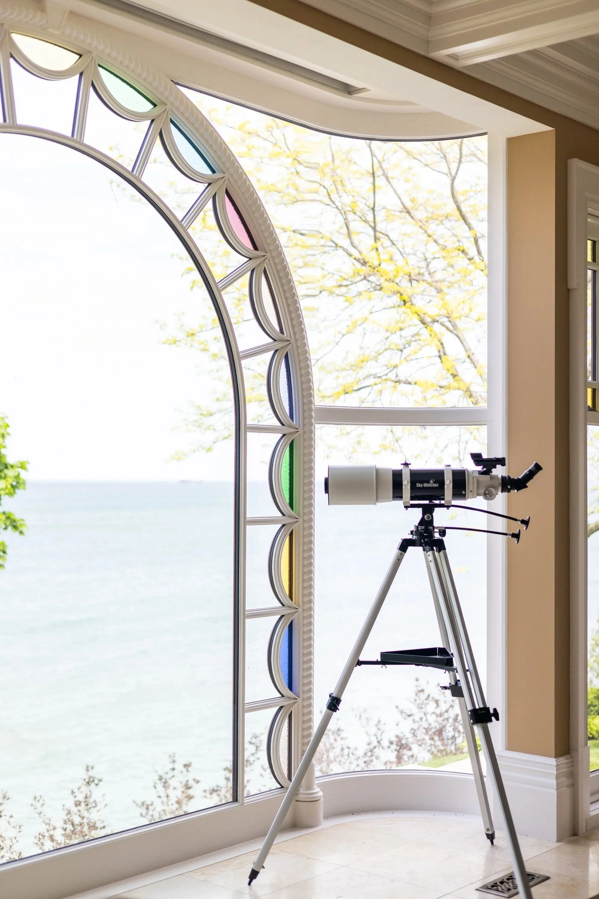 A telescope positioned inside a room near a large bay window with a scenic view of water and trees outside.